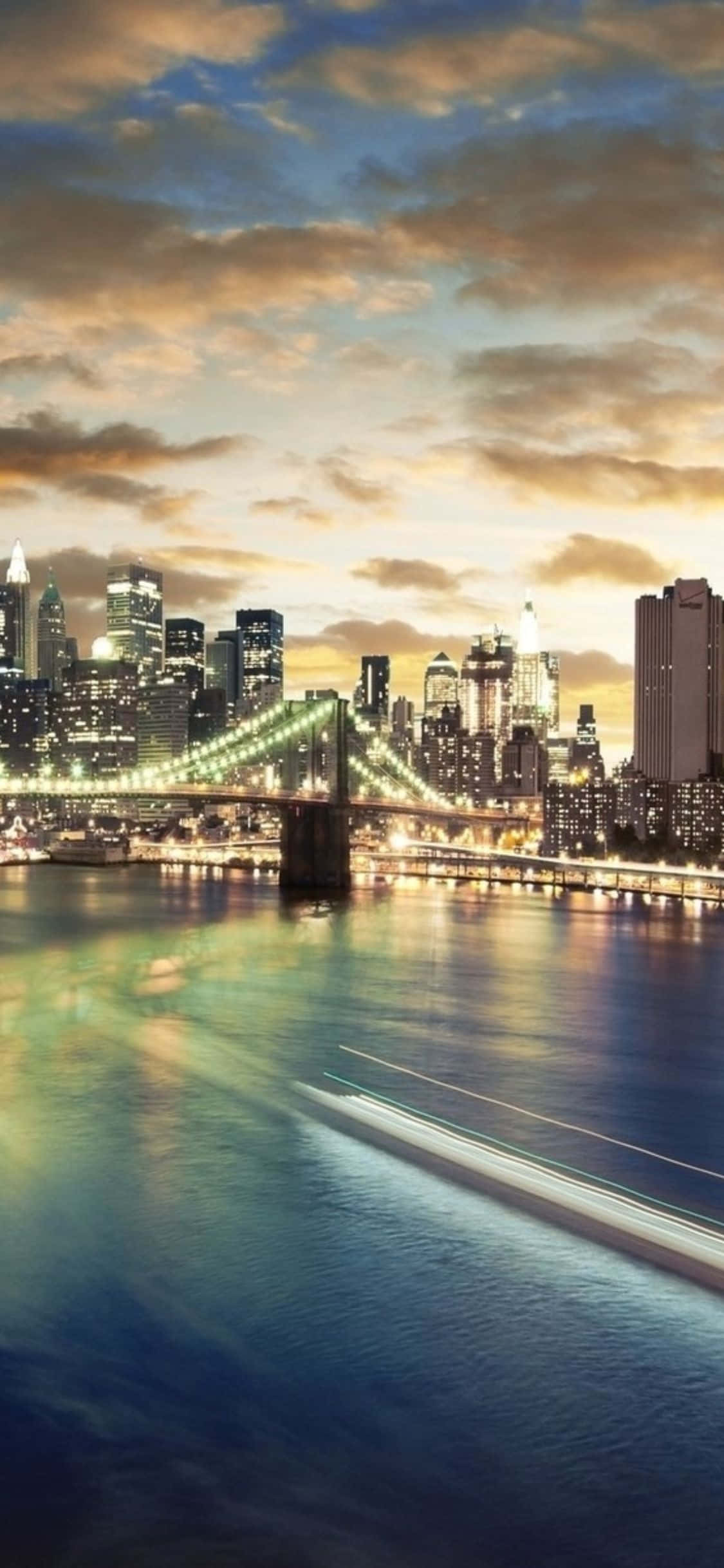 iPhone Xs Ny City 桌布 壁紙 — 布魯克林大橋和紐約天際線的壯麗黃昏景色，閃爍的城市燈光倒映在下方水面上。