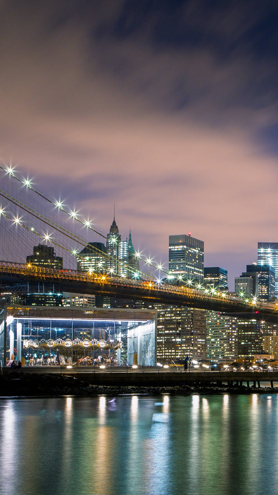 new york city iPhone x 桌布 壁紙 — 布魯克林大橋在夜晚閃耀，星芒燈光映照水面，反射城市摩天大樓，天空呈現柔和的粉紅與紫色。