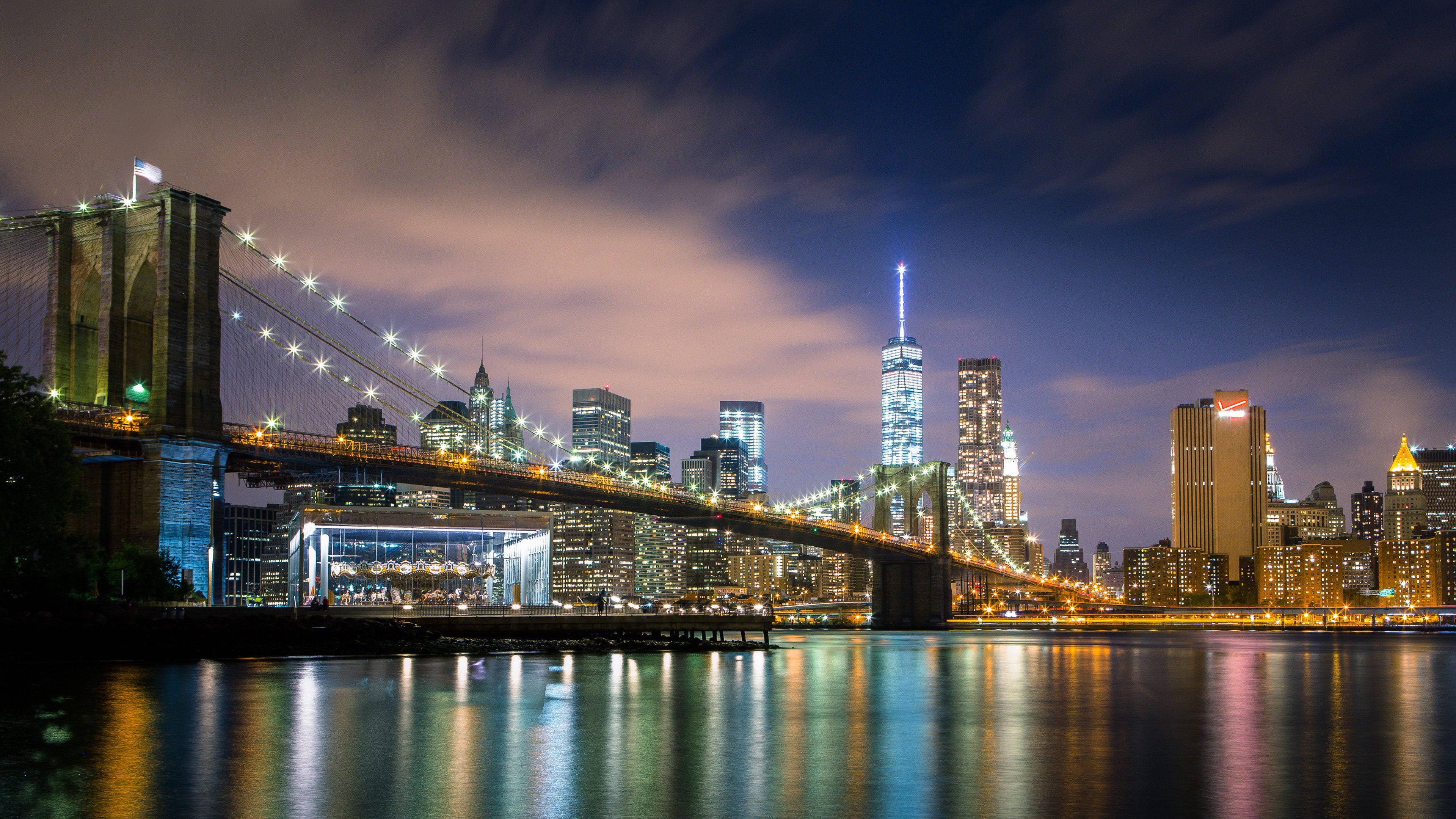 New York 桌布 壁紙 — 布魯克林大橋在夜燈下閃耀，燈光倒映在平靜的水面上，背景是曼哈頓的天際線。