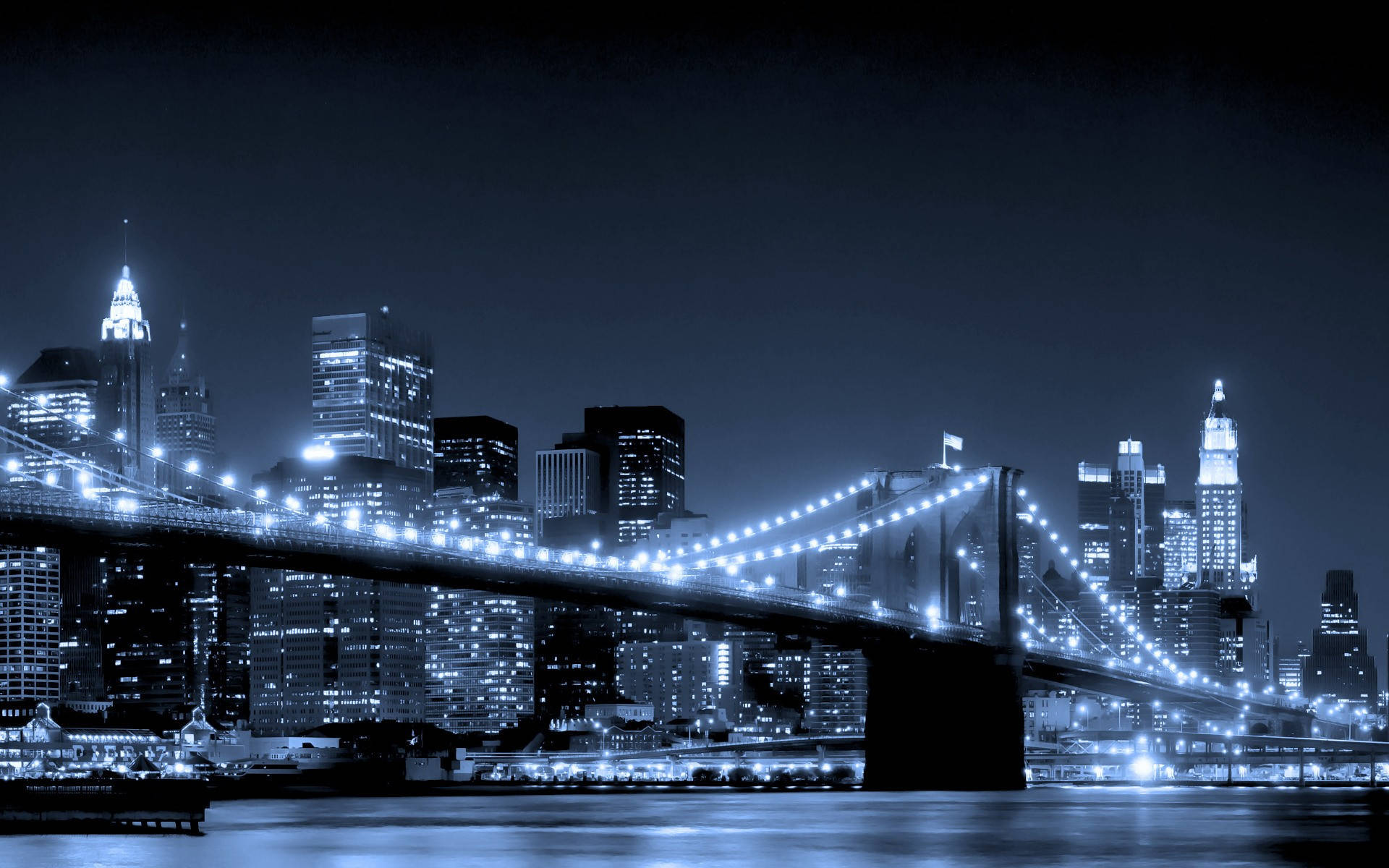 Brooklyn Bridge 桌布 壁紙 — 布魯克林大橋在夜間被發光的燈光照亮，燈光在黑暗的城市天際線背景下反射在水面上。