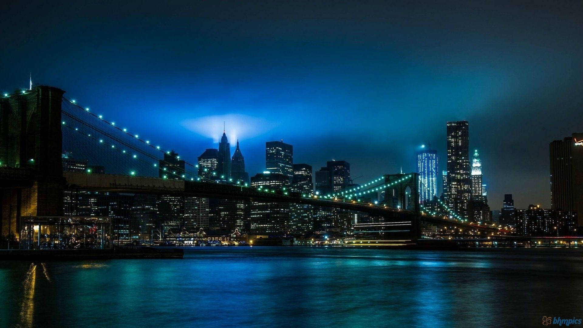 New York 桌布 壁紙 — 布魯克林大橋在深藍色天空下，夜晚被閃爍的燈光照亮，燈光倒映在水面上。