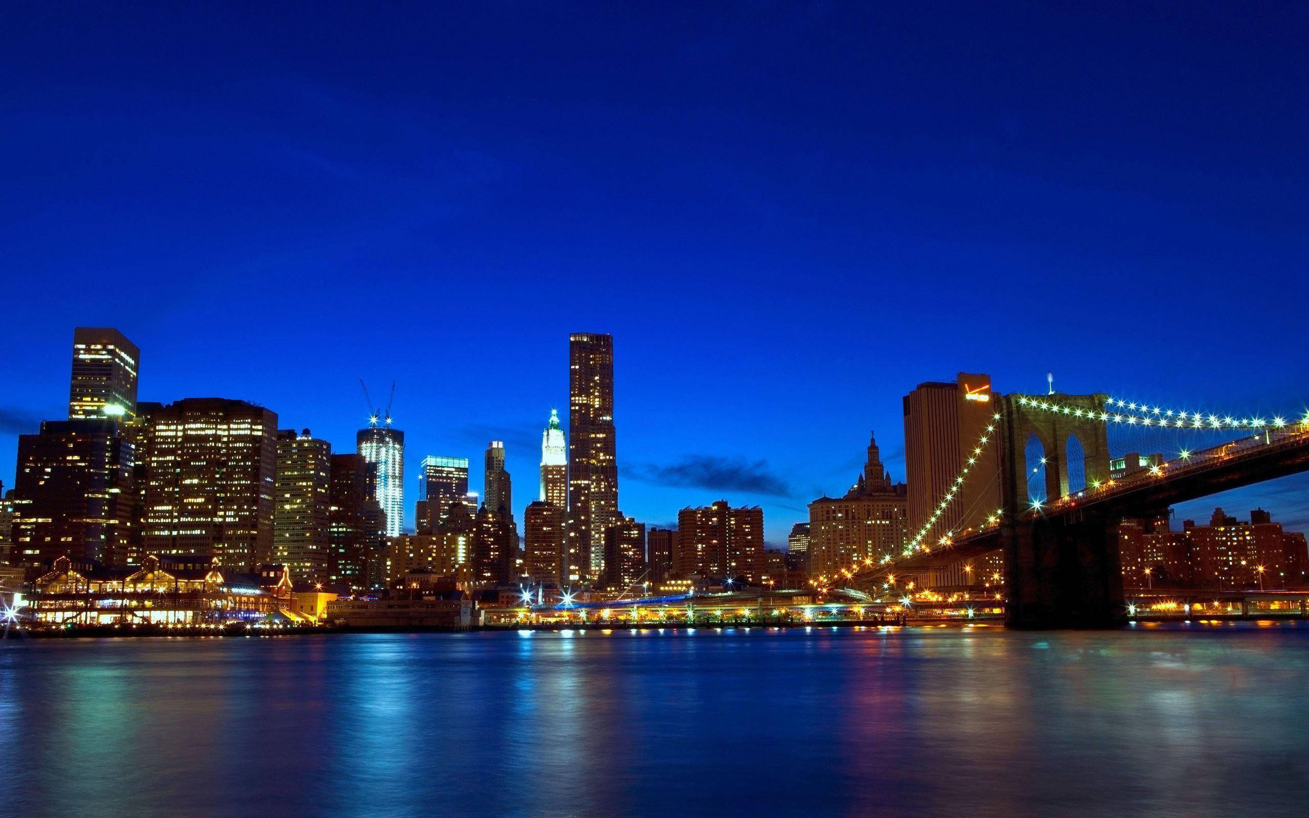 New York City Desktop 桌布 壁紙 — 布魯克林大橋在深藍色黃昏天空的映襯下，散發著溫暖的光芒，下方是倒映著燈光的東河和曼哈頓天際線。