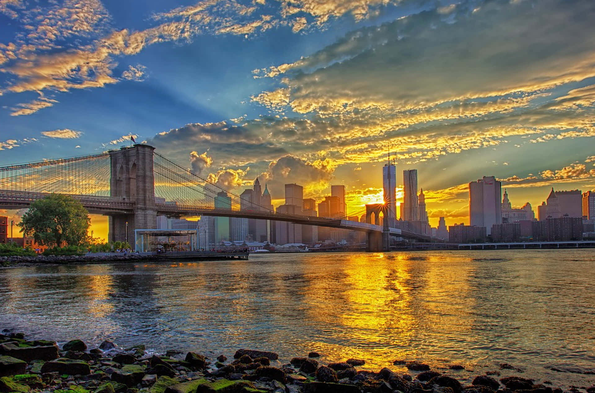 Nyc 桌布 壁紙 — 布魯克林大橋在金色日落時橫跨河流，摩天大樓在戲劇性的雲層下倒映在水中。