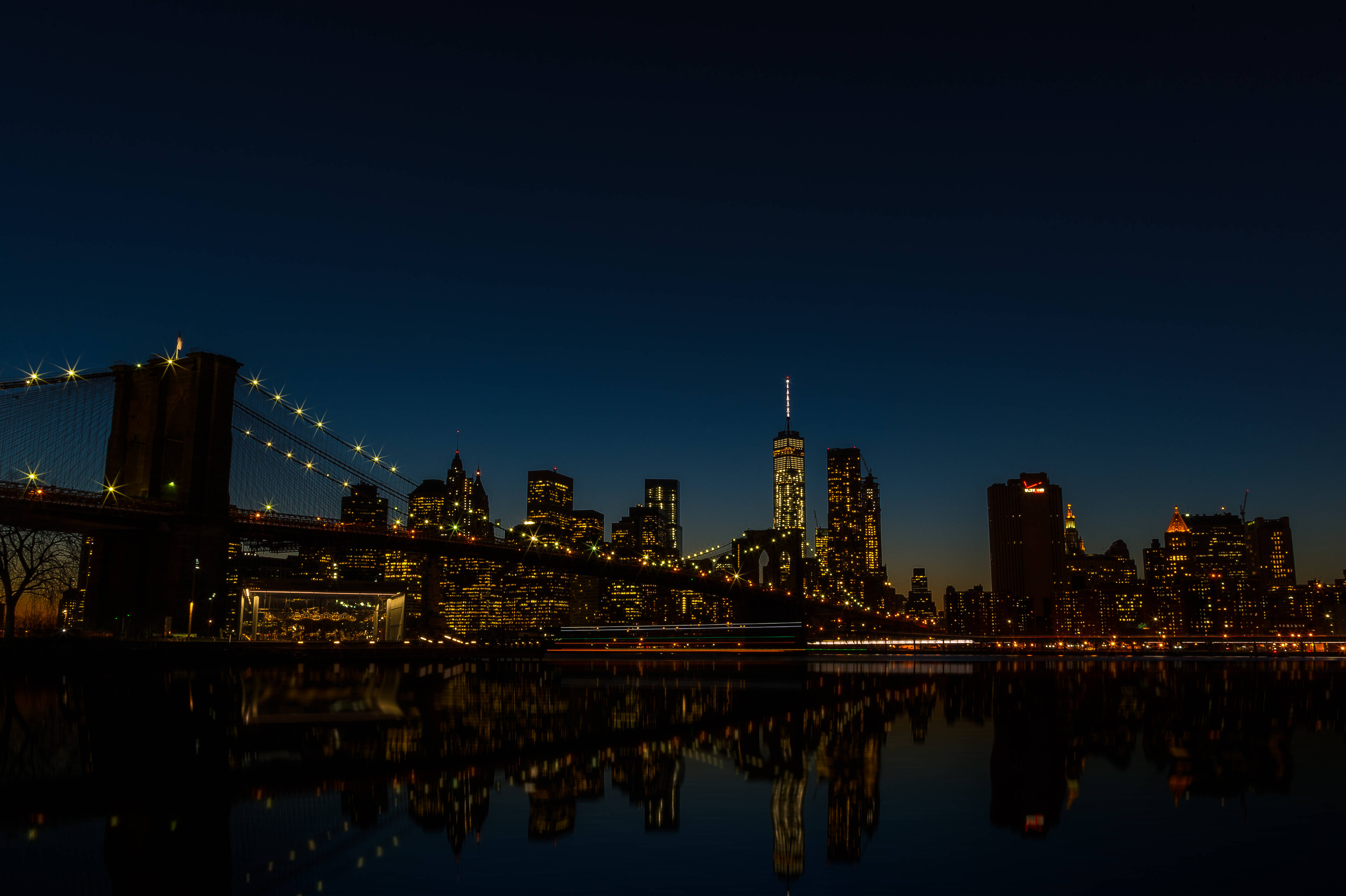 New York 桌布 壁紙 — 布魯克林大橋在黃昏時分閃耀，城市燈光倒映在平靜的水面上，襯托著深藍色的天空。