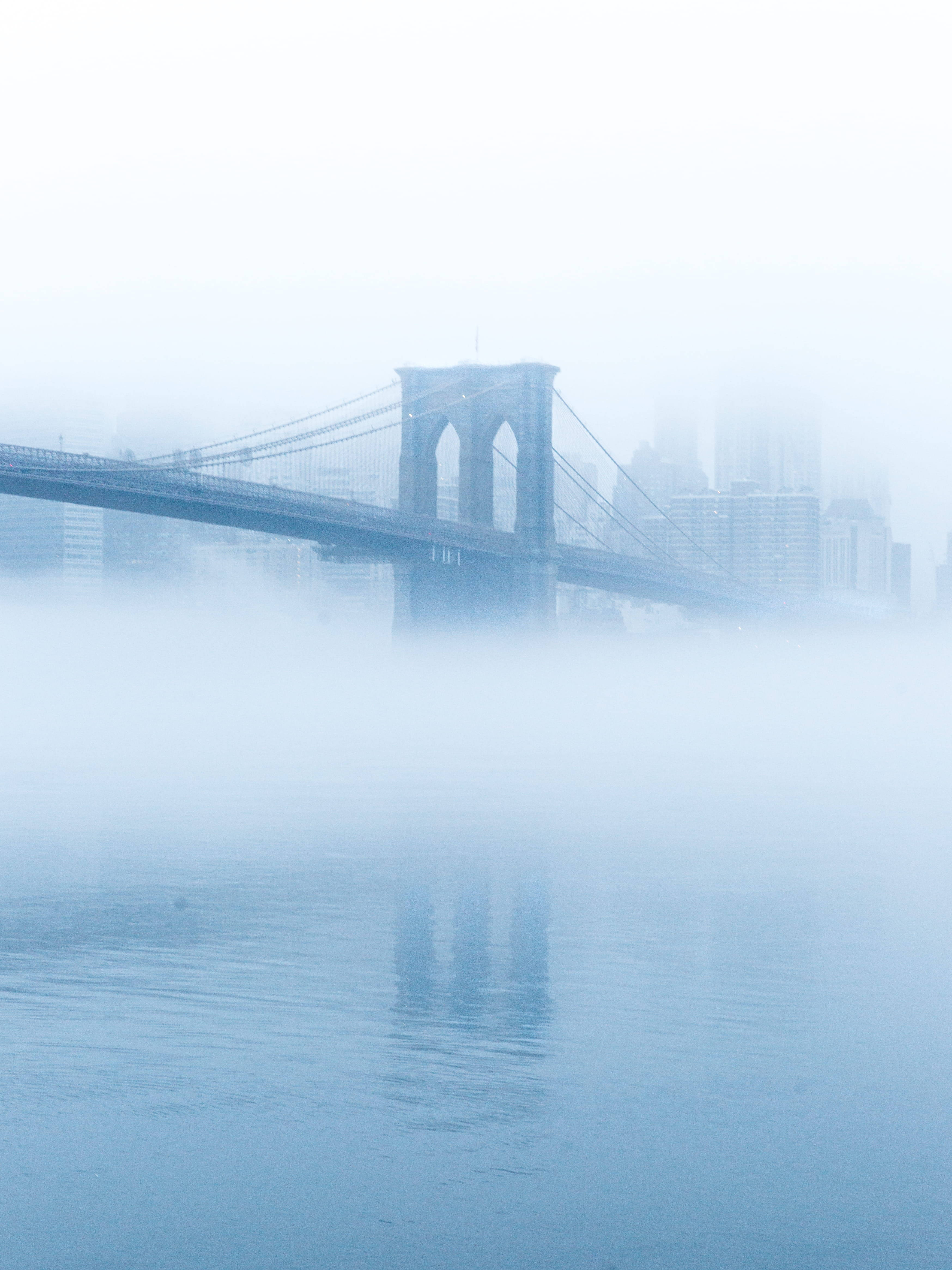 New York 桌布 壁紙 — 布魯克林大橋從濃霧中浮現，橫跨平靜的水面，城市天際線在霧濛濛的背景中隱約可見。
