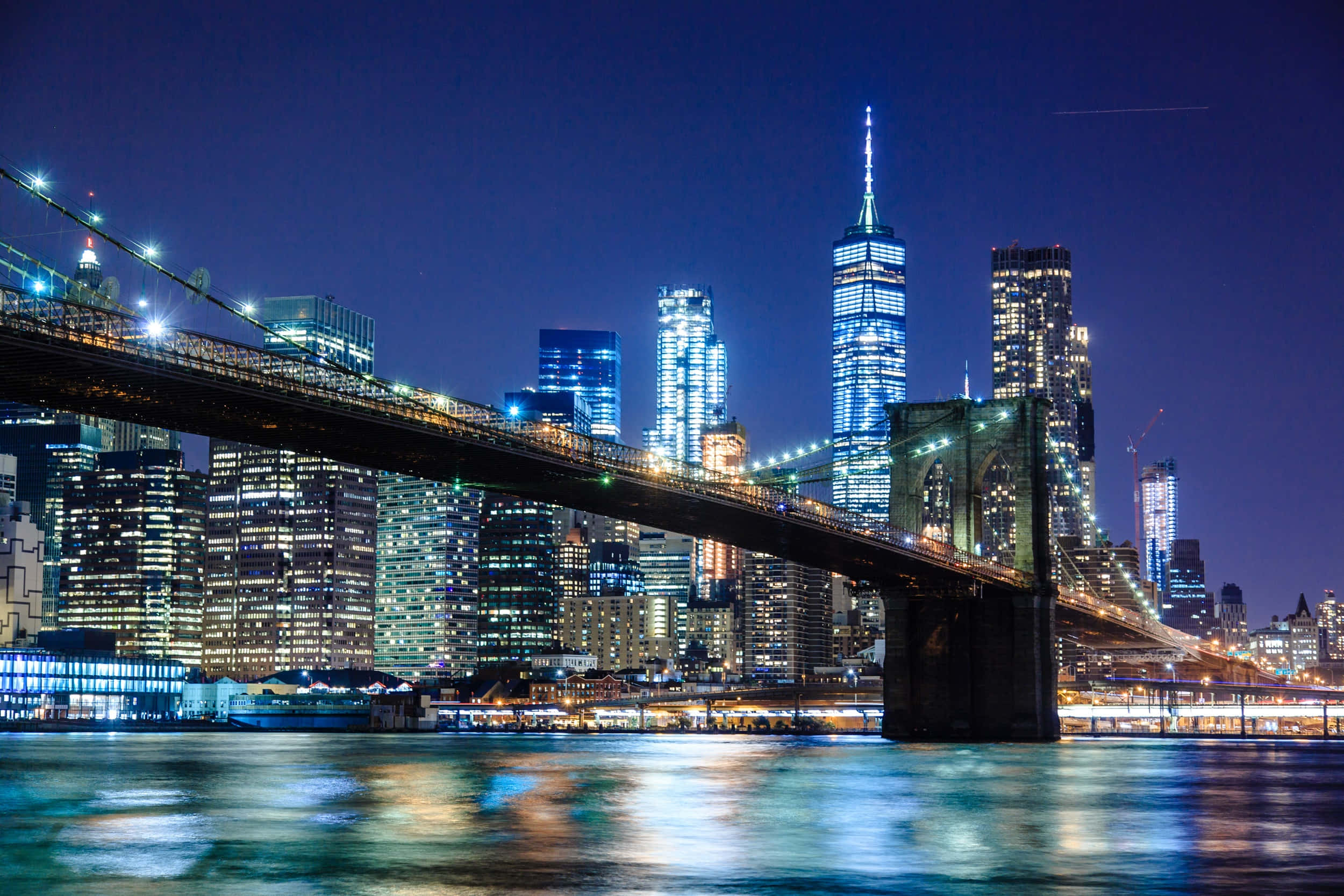 Brooklyn Bridge 桌布 壁紙 — 布魯克林大橋橫跨一條發光的河流，背景是深藍色的夜空和燈火通明的摩天大樓。