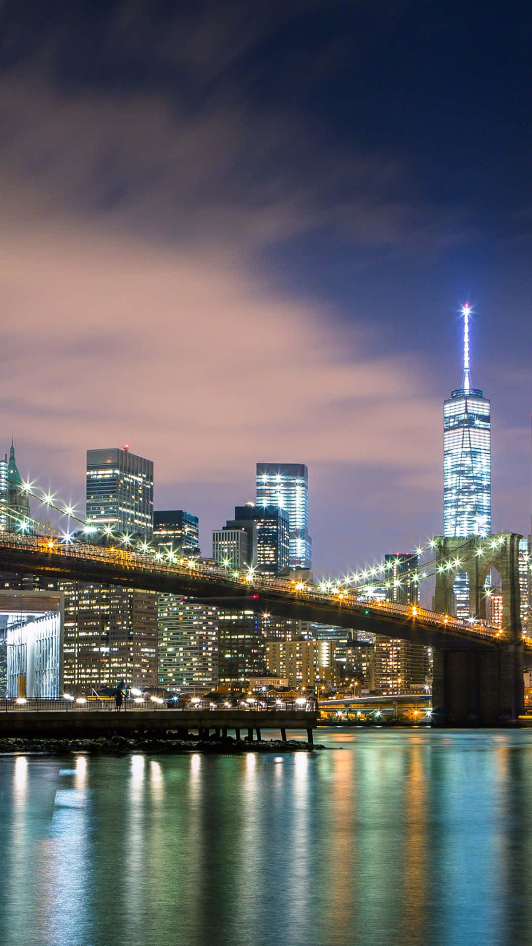 Android Ny City 桌布 壁紙 — 布魯克林大橋橫跨平靜的河流，在暮色天空下，閃爍的城市燈光倒映在水面上，背景中世界貿易中心一號大樓高聳入雲。