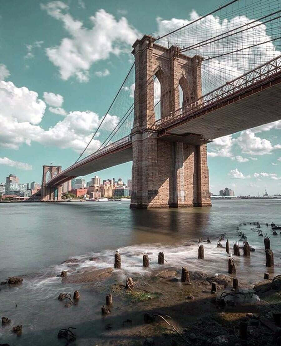 bridge 桌布 壁紙 — 布魯克林大橋橫跨東河，天空明亮，背景是城市天際線，前景是風化的樁柱。