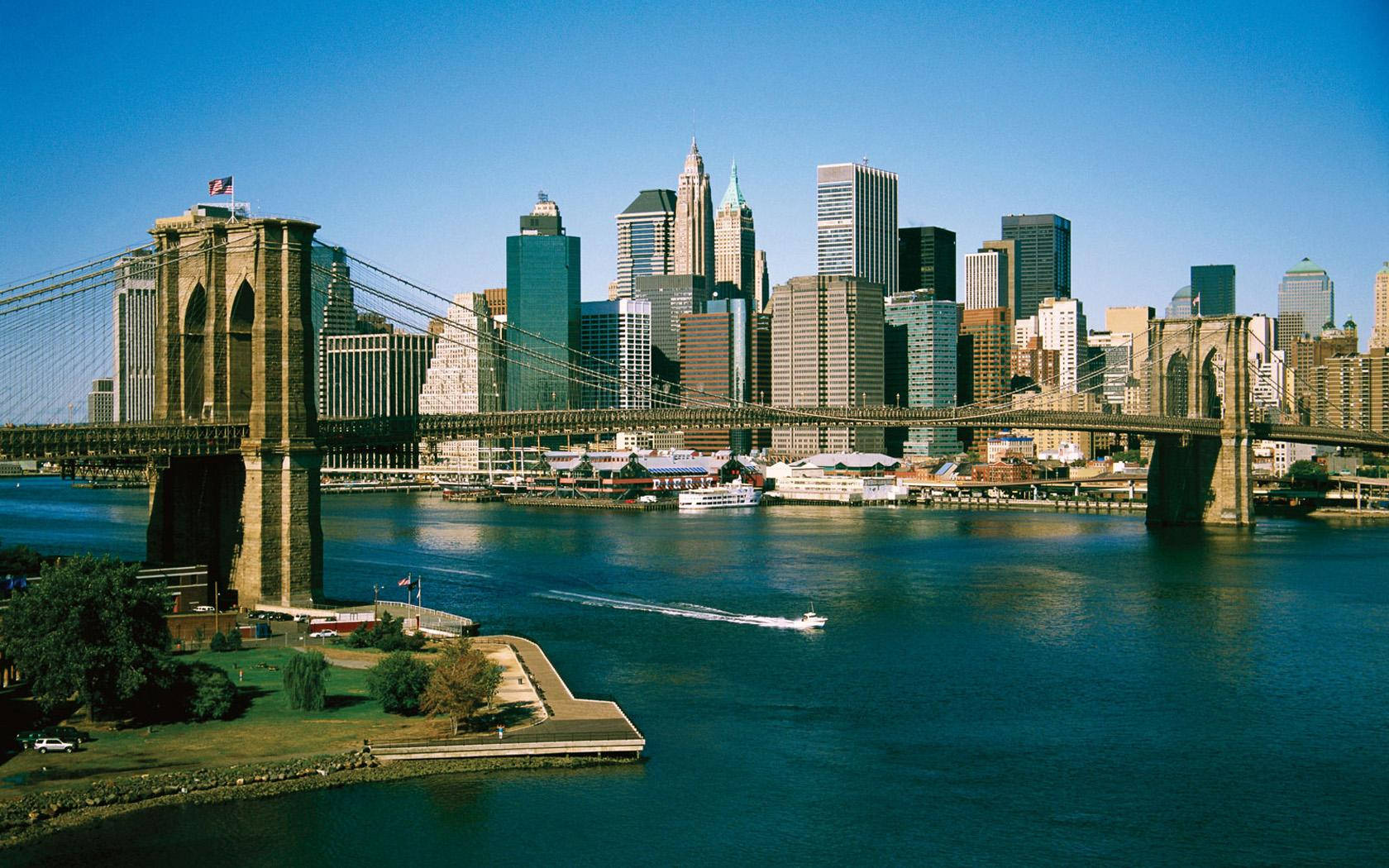 Brooklyn Bridge 桌布 壁紙 — 布魯克林大橋橫跨深藍色的河流，城市天際線由玻璃和鋼鐵塔樓構成，在清澈的藍天下。