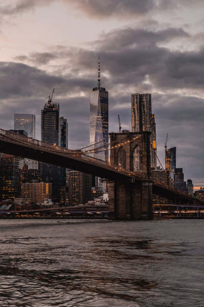 bridge 桌布 壁紙 — 布魯克林大橋橫跨黑暗的河流，背景是陰鬱的黃昏天空和燈火通明的摩天大樓。