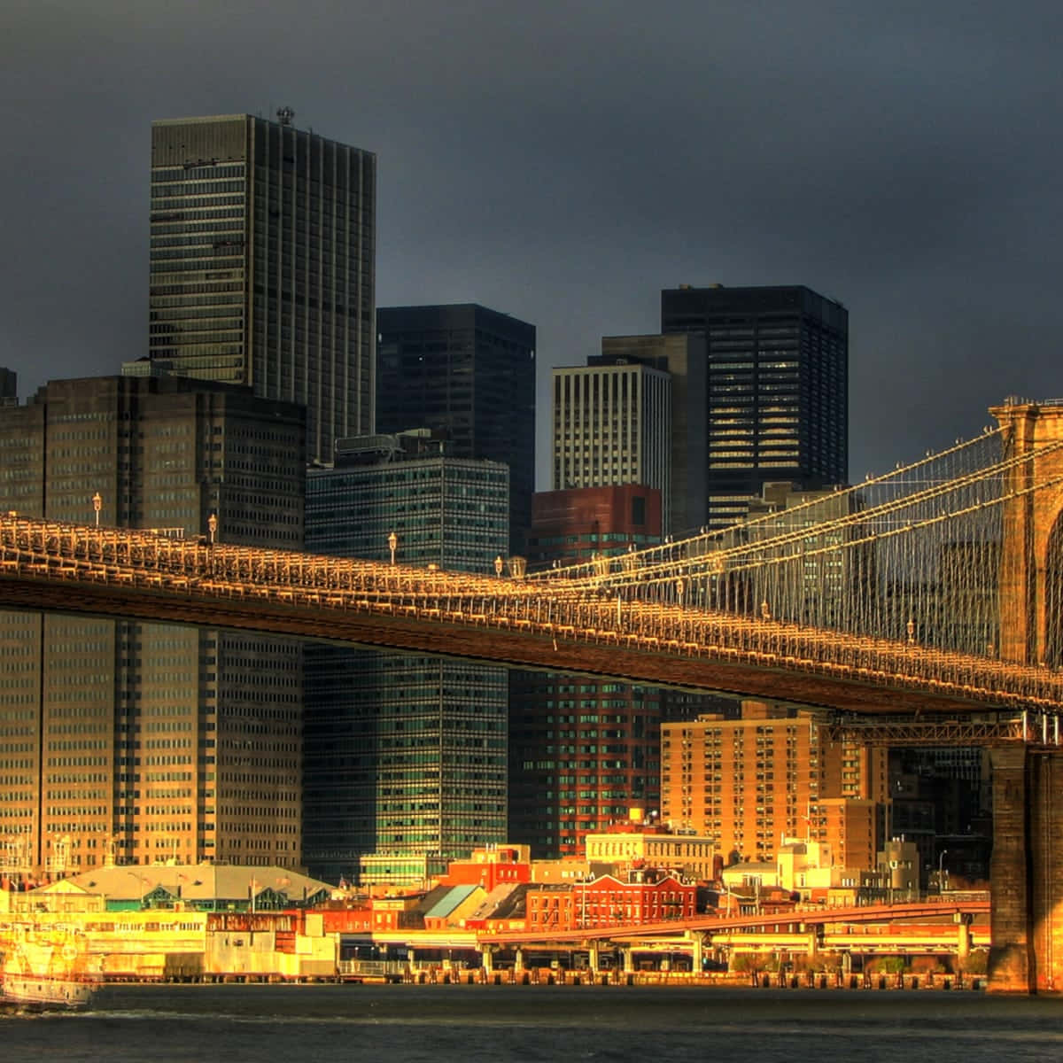 Nyc 桌布 壁紙 — 布魯克林大橋沐浴在金色日落光芒中，橫跨河流，背後是高聳的摩天大樓，天空陰沉。