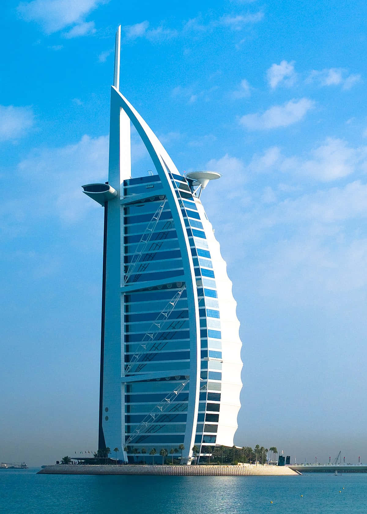 Building 桌布 壁紙 — 帆船形狀的 Burj Al Arab Jumeirah 飯店在蔚藍天空和零星白雲下閃耀，玻璃外牆將陽光反射在平靜的碧綠水面上。