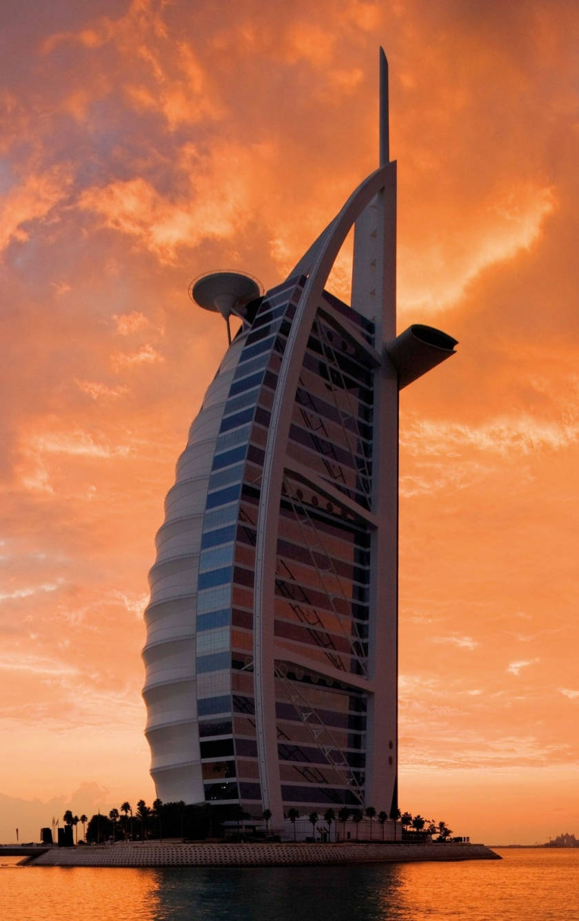 日落 桌布 壁紙 — 帆船形狀的帆船酒店（Burj Al Arab）雄偉地矗立在寧靜水面上倒映的火熱橙色日落天空前。