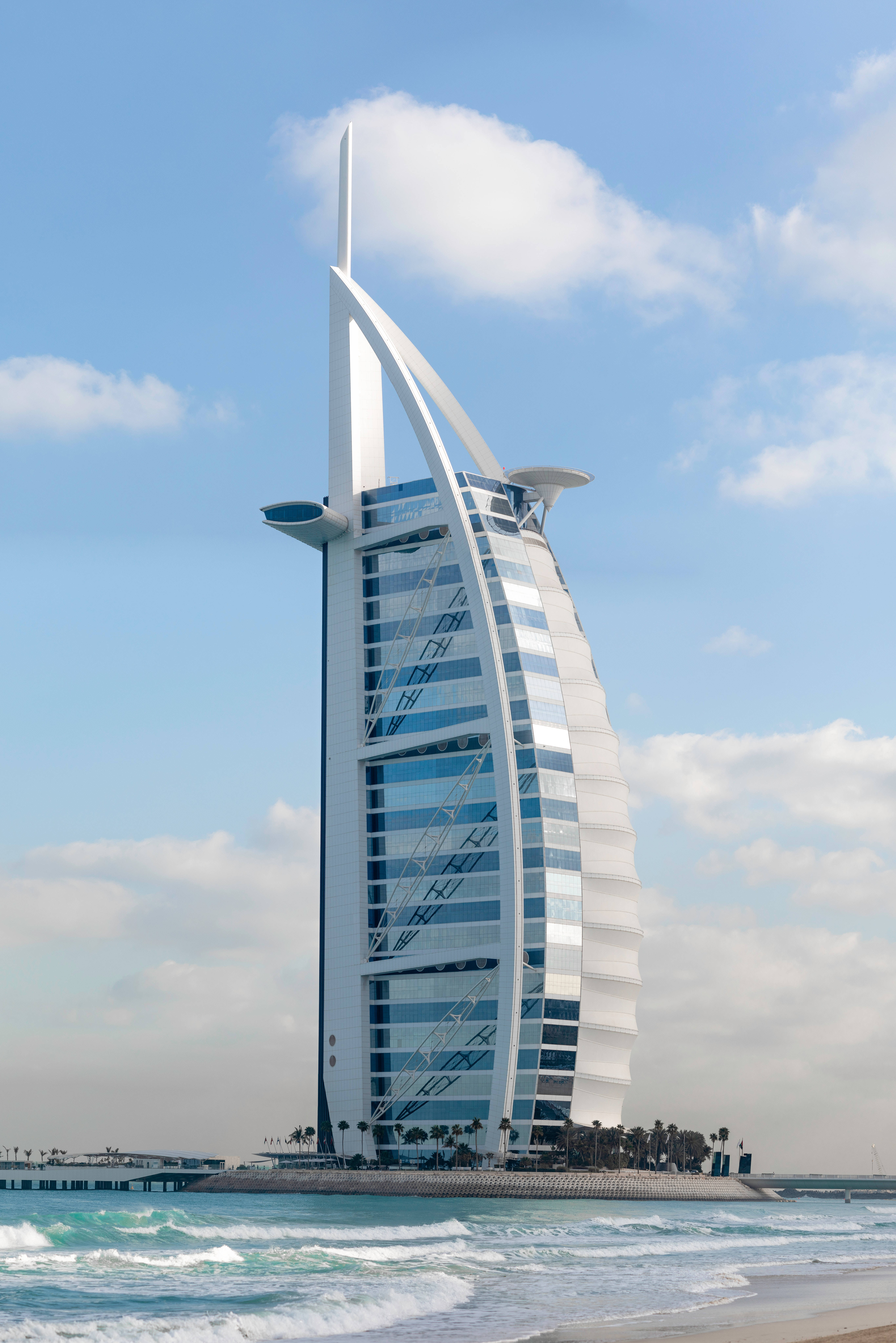 iPhone 桌布 壁紙 — 帆船形狀的帆船飯店 Burj Al Arab Jumeirah 高聳在明亮的藍天下，有散落的雲朵和附近翻騰的碧綠海浪。