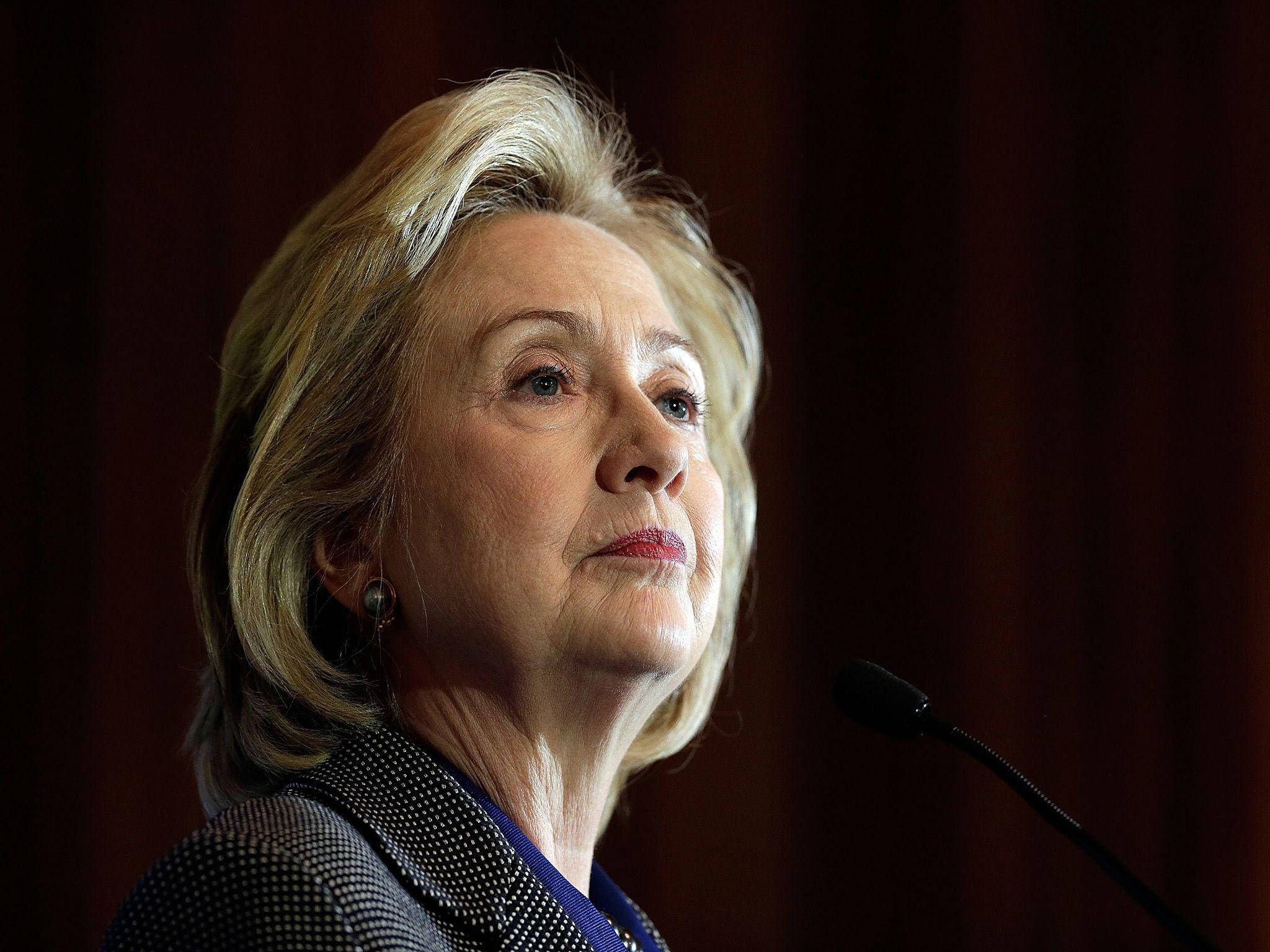 hillary clinton 桌布 壁紙 — 希拉蕊·柯林頓側面站在講台上，拿著麥克風，背景是深紅色窗簾。