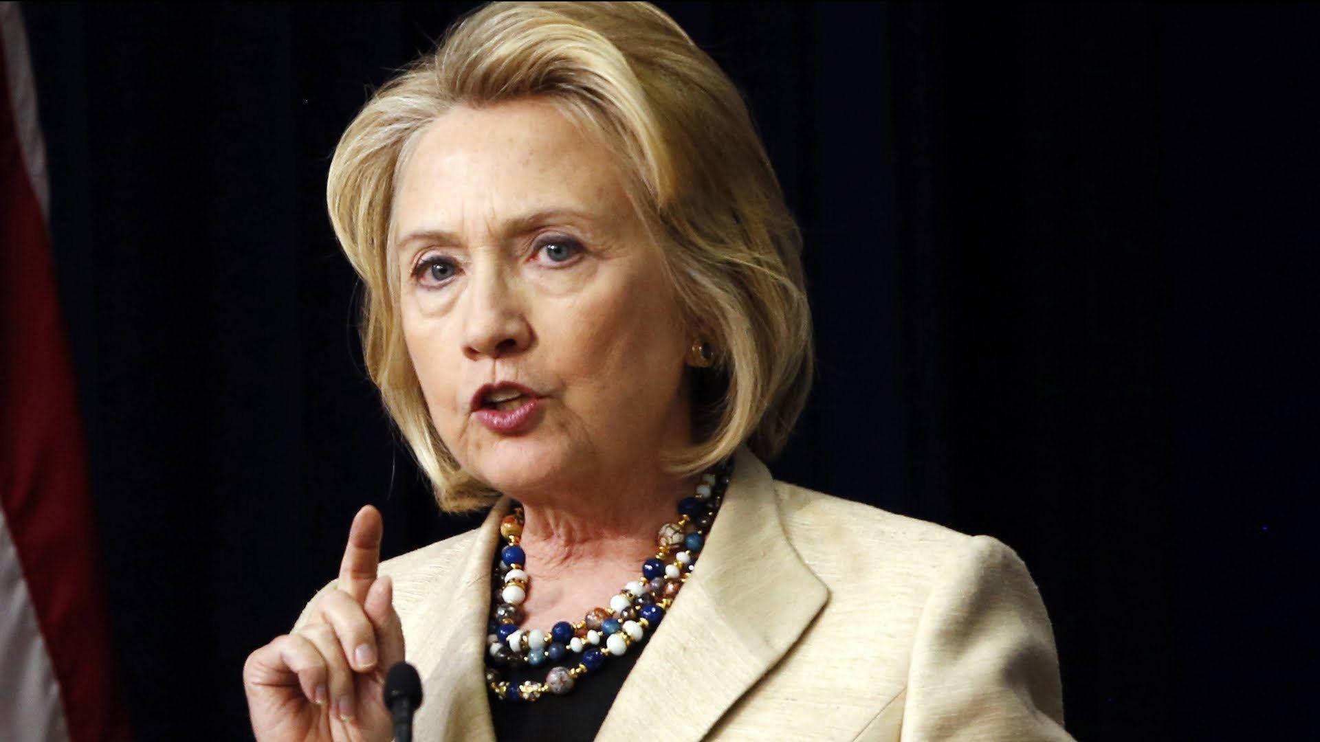 hillary clinton 桌布 壁紙 — 希拉蕊·柯林頓在深色背景下，身穿米色西裝外套和珠鏈，手指指點，激烈地說話。