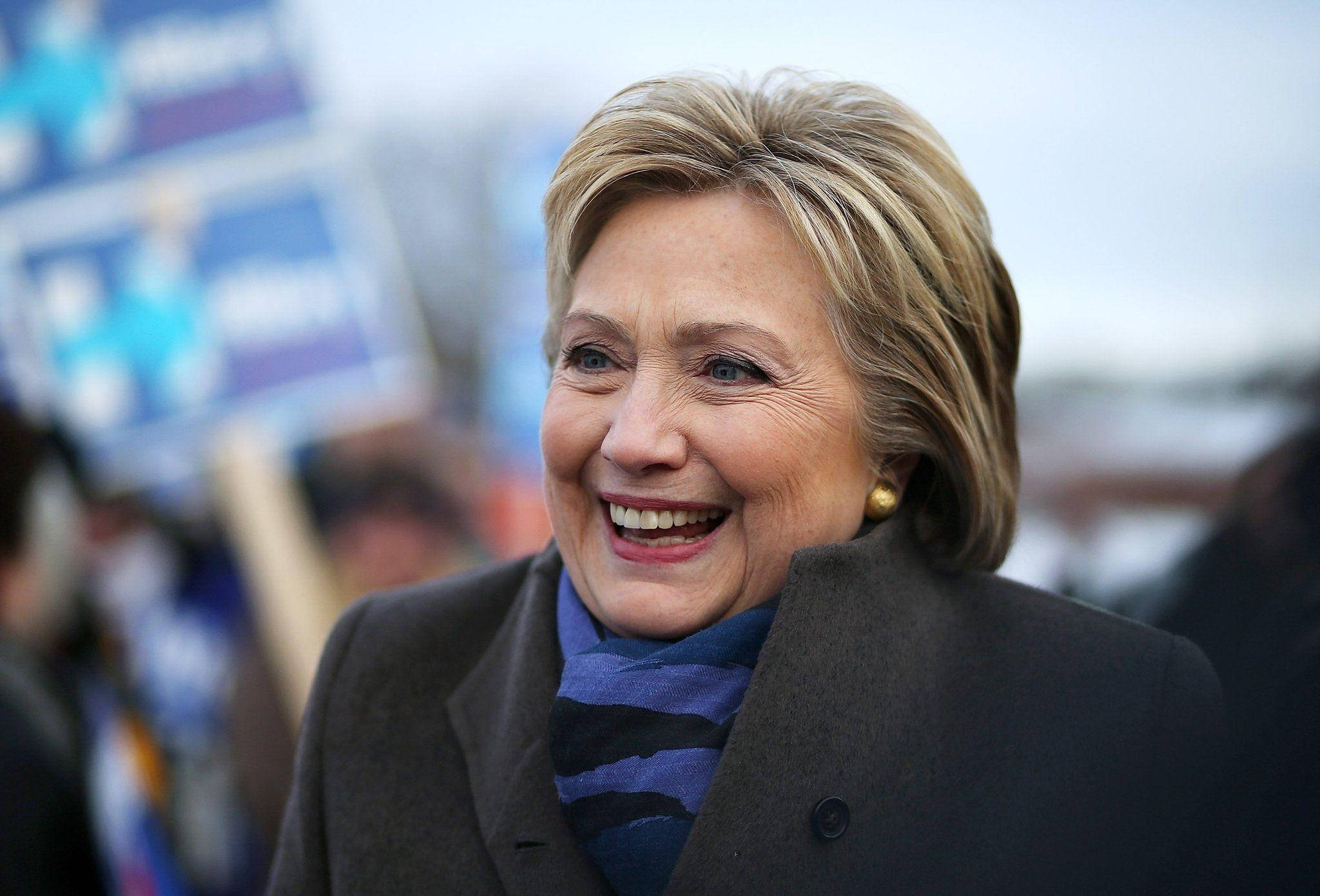 Smile 桌布 壁紙 — 希拉蕊·柯林頓在競選集會上溫暖微笑，背後有模糊的支持者和標語牌。