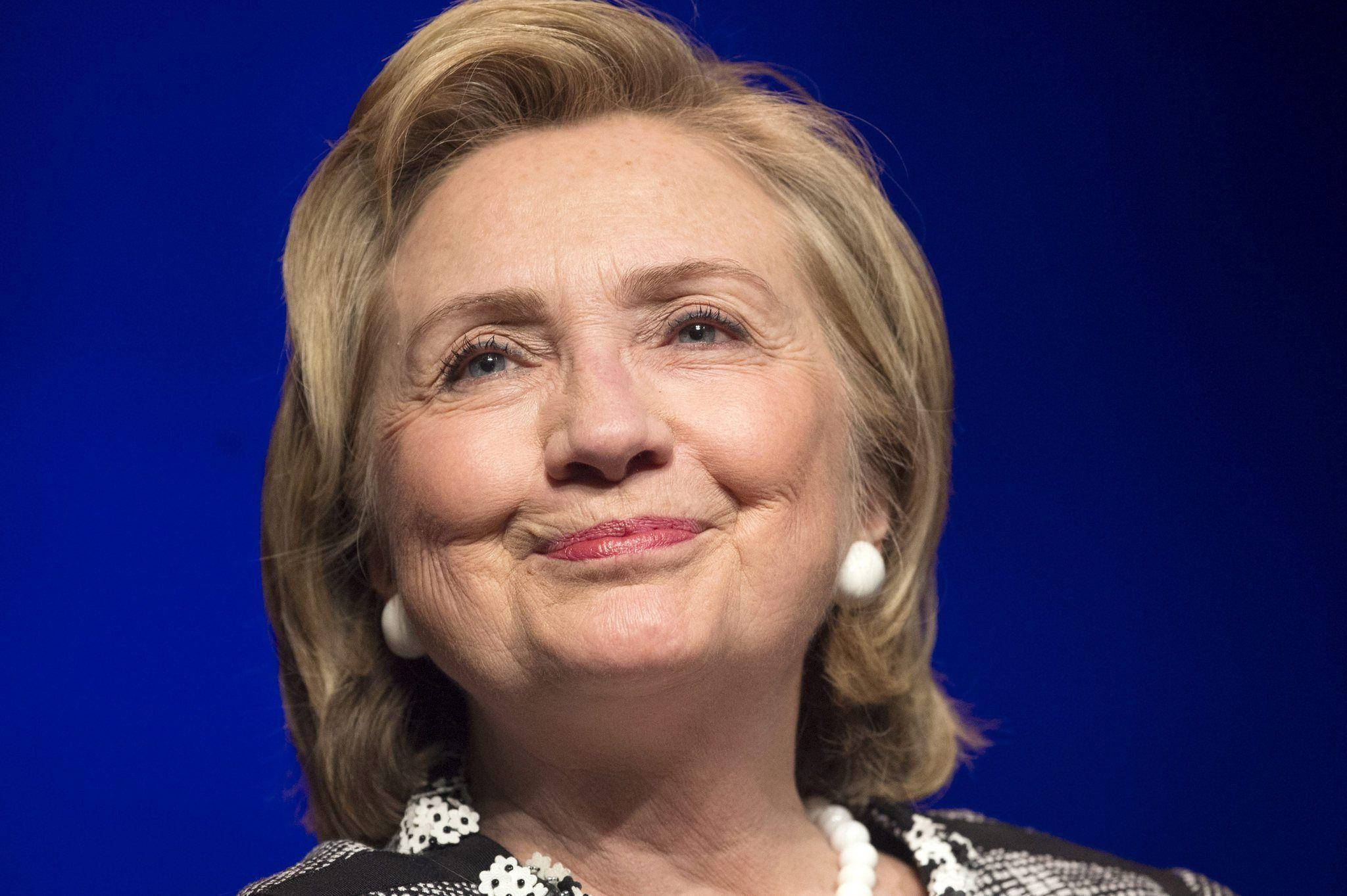 hillary clinton 桌布 壁紙 — 希拉蕊·柯林頓在純藍色背景前溫暖微笑，戴著珍珠耳環和圖案上衣。