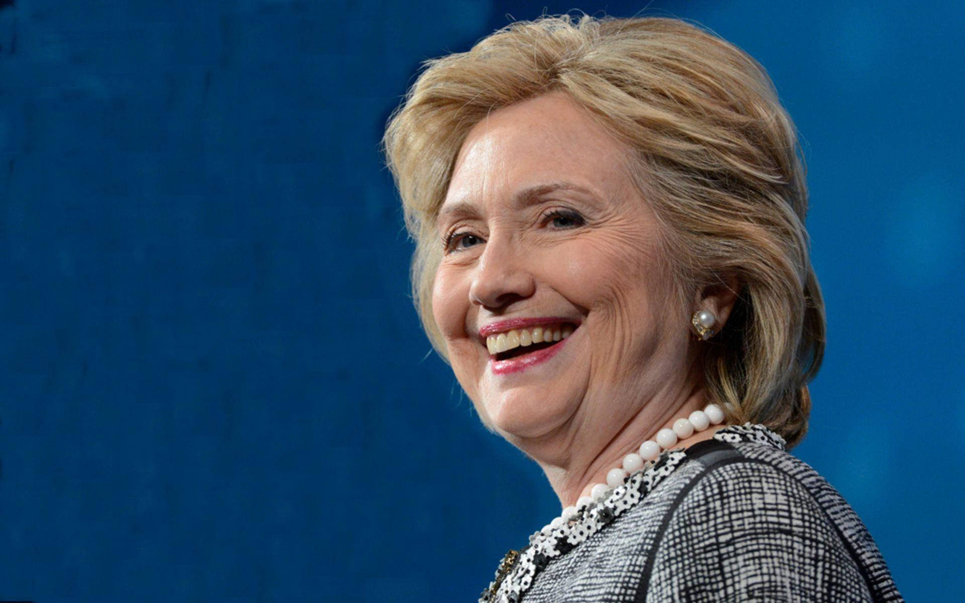 hillary clinton 桌布 壁紙 — 希拉蕊·柯林頓在純藍色背景前的特寫肖像中溫暖微笑，佩戴珍珠和圖案外套。