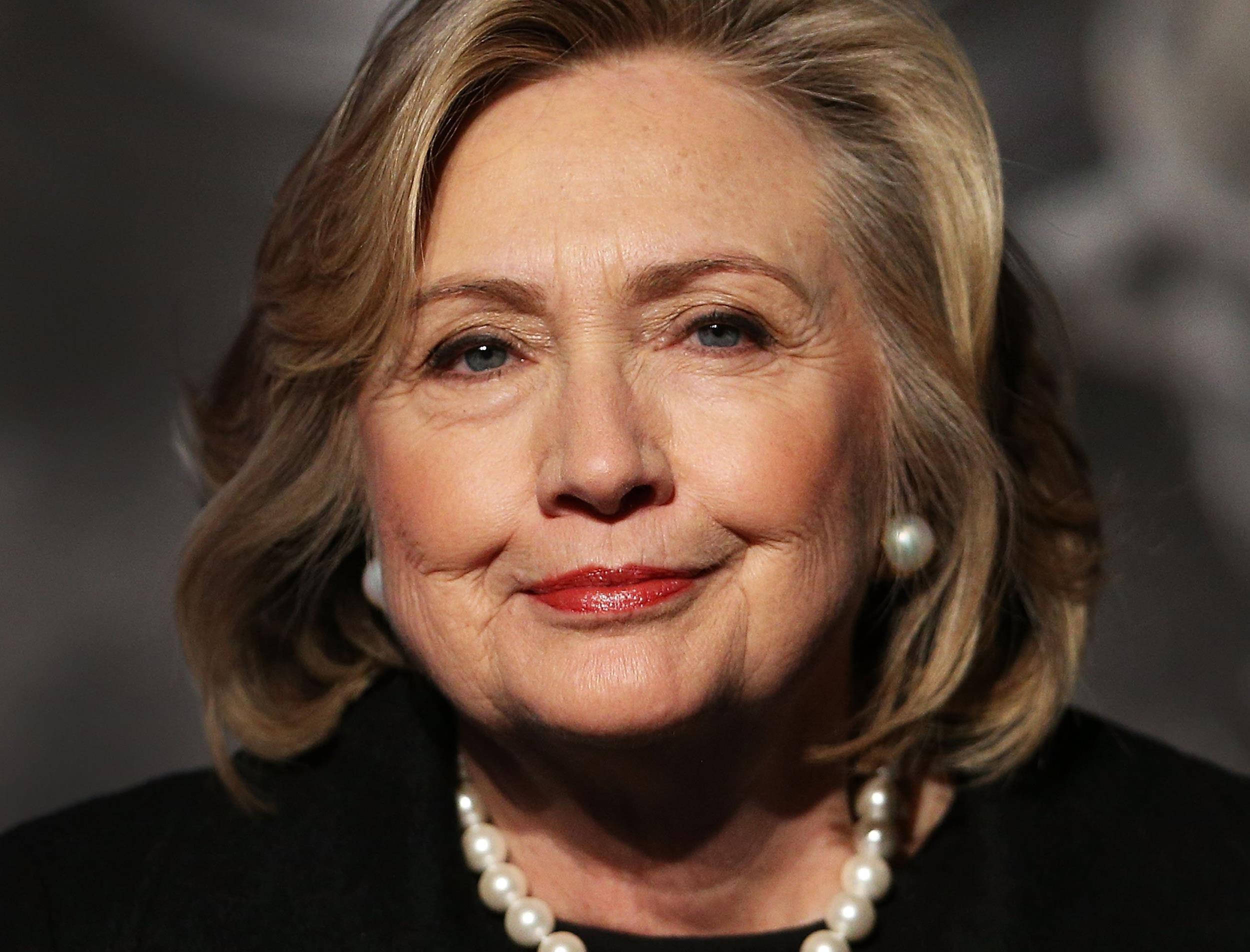 hillary clinton 桌布 壁紙 — 希拉蕊·柯林頓的近距離肖像，戴著珍珠耳環和項鍊，在模糊的灰色背景下溫暖地微笑。
