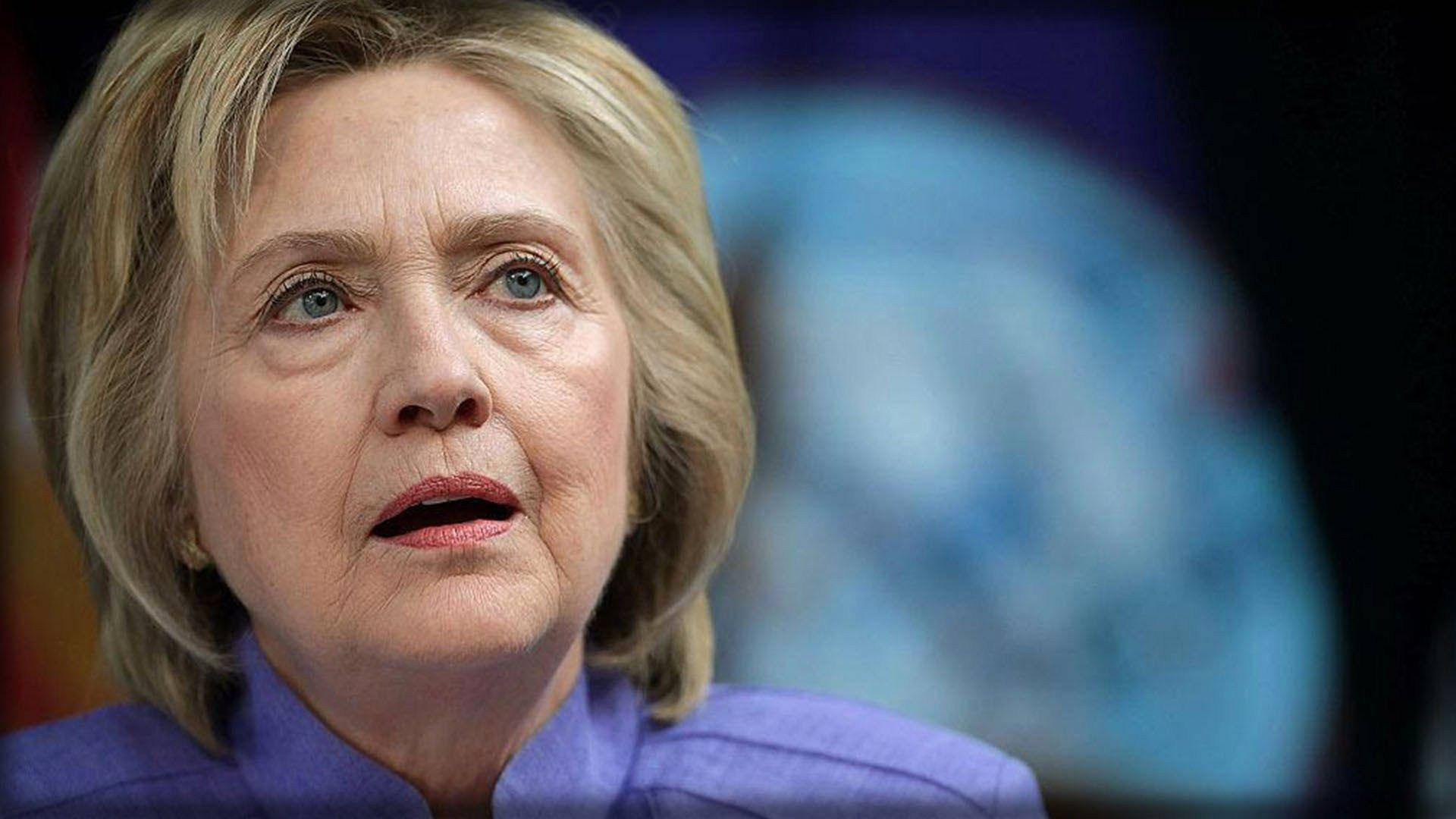 hillary clinton 桌布 壁紙 — 希拉蕊·柯林頓身穿尖銳的紫色西裝，在模糊的藍色地球背景下，表情嚴肅地向上凝視。