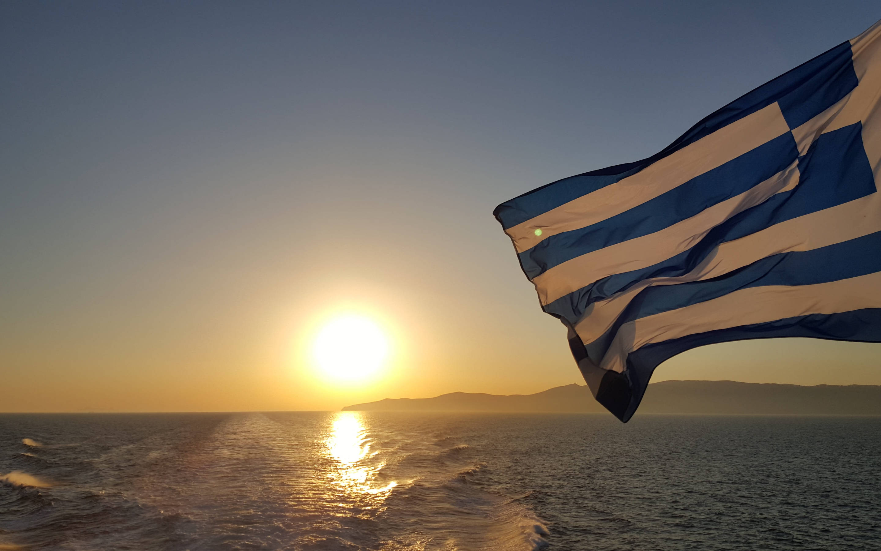greek 桌布 壁紙 — 希臘國旗在平靜海面上的金色日落背景下自豪地飄揚，遠方有山丘。