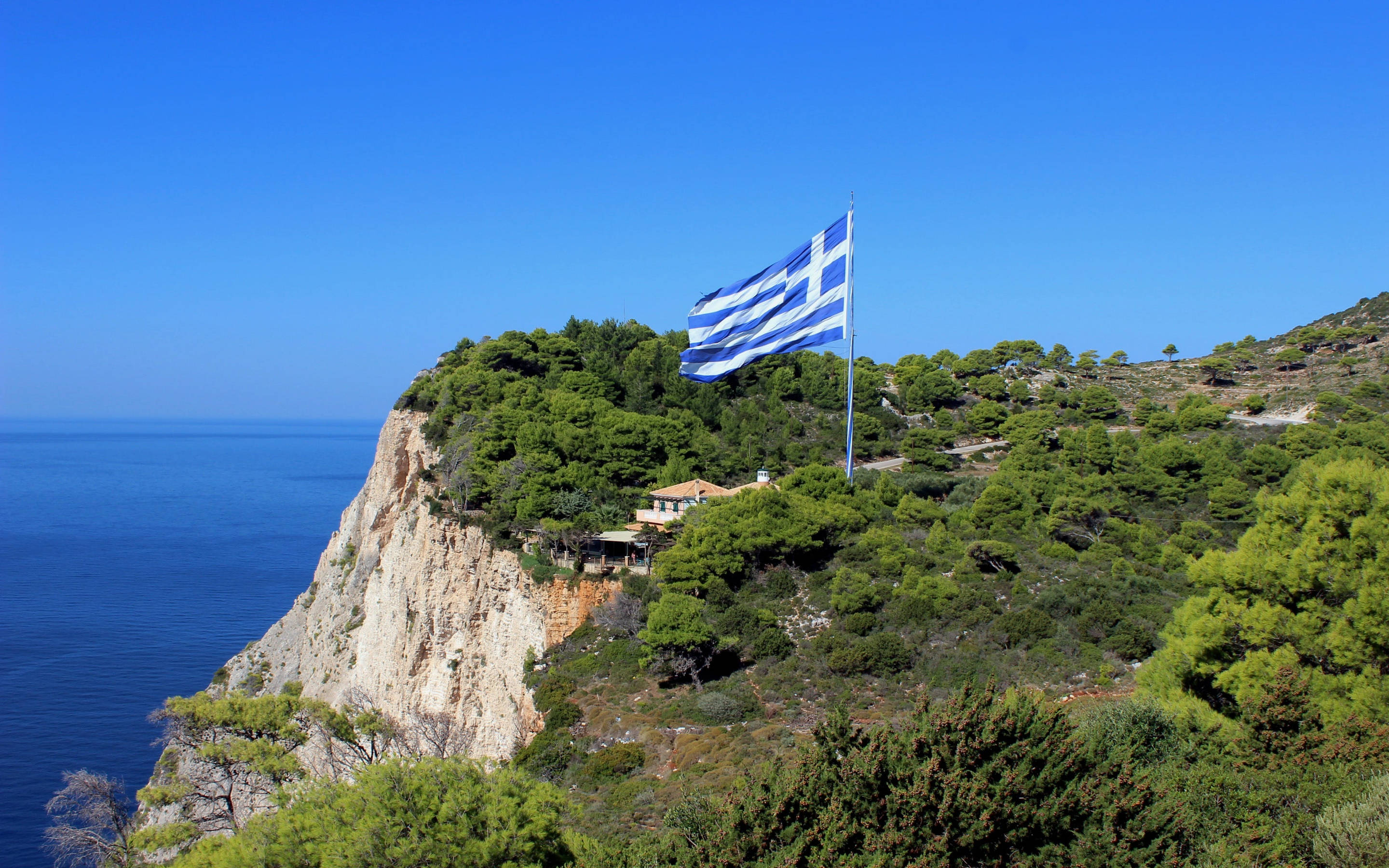 greek 桌布 壁紙 — 希臘國旗在陽光明媚的懸崖頂上自豪地飄揚，俯瞰著深藍色的愛琴海和下方鬱鬱蔥蔥的綠色植物。