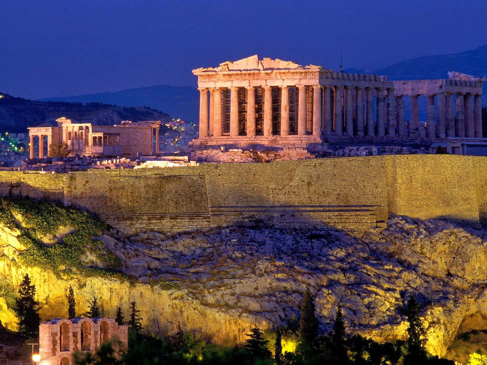 greek 桌布 壁紙 — 帕德嫩神廟和衛城在暮色燈光下溫暖地發光，映襯著深藍色的天空，遠處的城市燈光在下方閃爍。