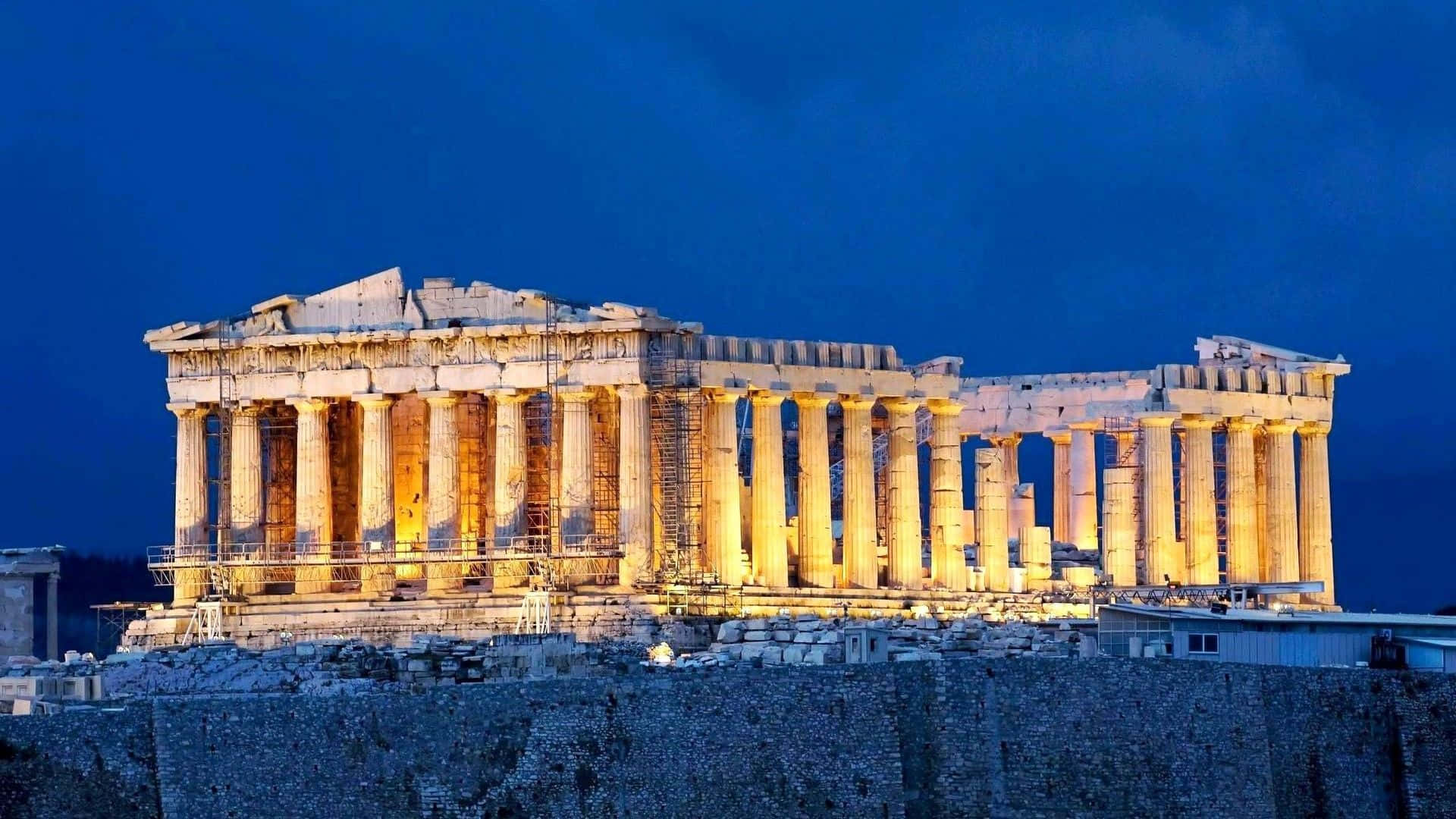 greek 桌布 壁紙 — 帕德嫩神廟在深藍色的暮色天空下被照亮，溫暖的金色燈光突顯其古老的柱子和石結構。