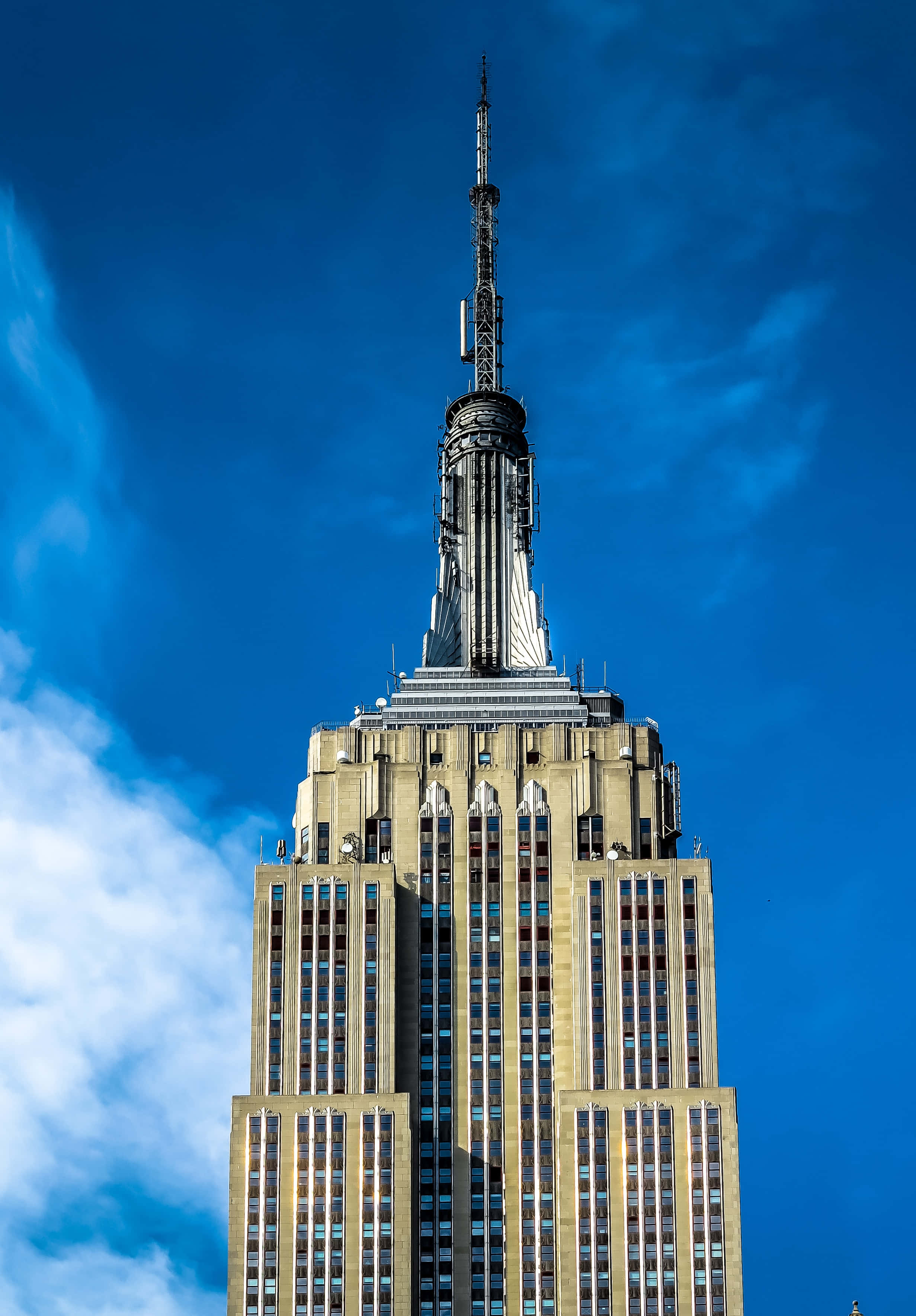 Empire State Building 桌布 壁紙 — 帝國大廈刺穿一片帶有縷縷白雲的鮮豔藍天，展示其標誌性的裝飾藝術風格尖頂和精緻的外牆。