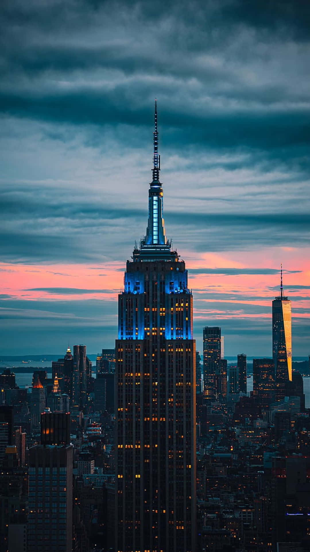 new york city iPhone x 桌布 壁紙 — 帝國大廈在佈滿粉紅色和深色雲彩的暮色天空中閃耀著藍光，下方是密集的紐約市天際線。