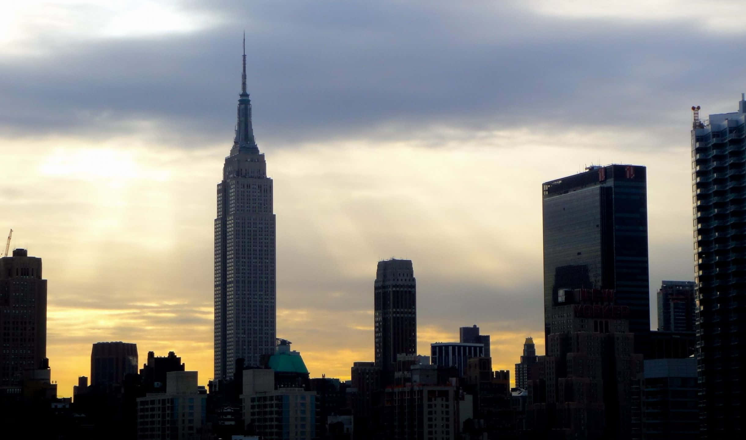 2440x1440 Empire State Building 桌布 壁紙 — 帝國大廈在壯麗的日落天空下高聳入雲，金色雲彩和城市天際線剪影交相輝映。