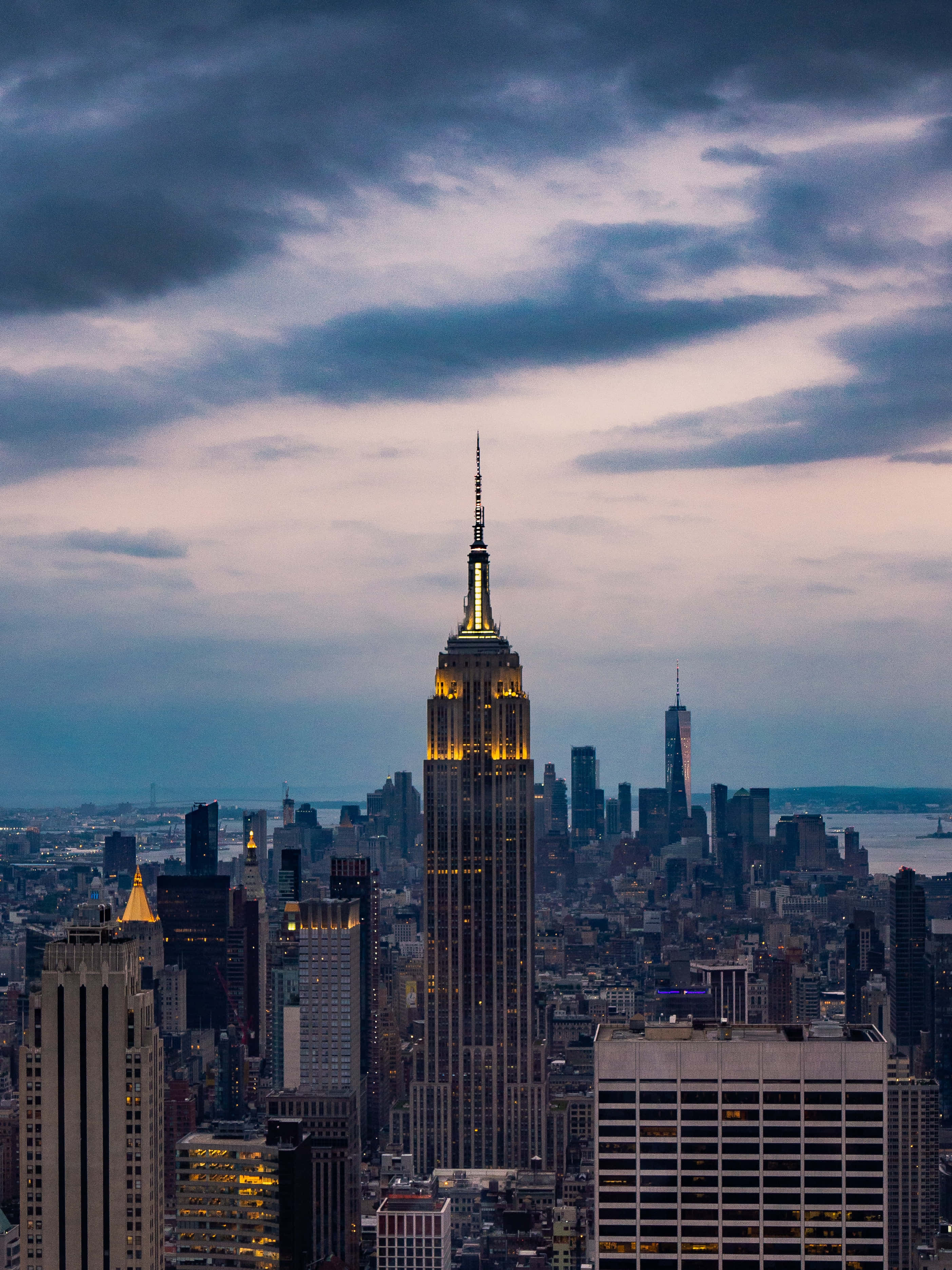 Empire State Building 桌布 壁紙 — 帝國大廈在憂鬱的暮色天空中閃耀著金色光芒，紐約市的天際線在其下方延伸。
