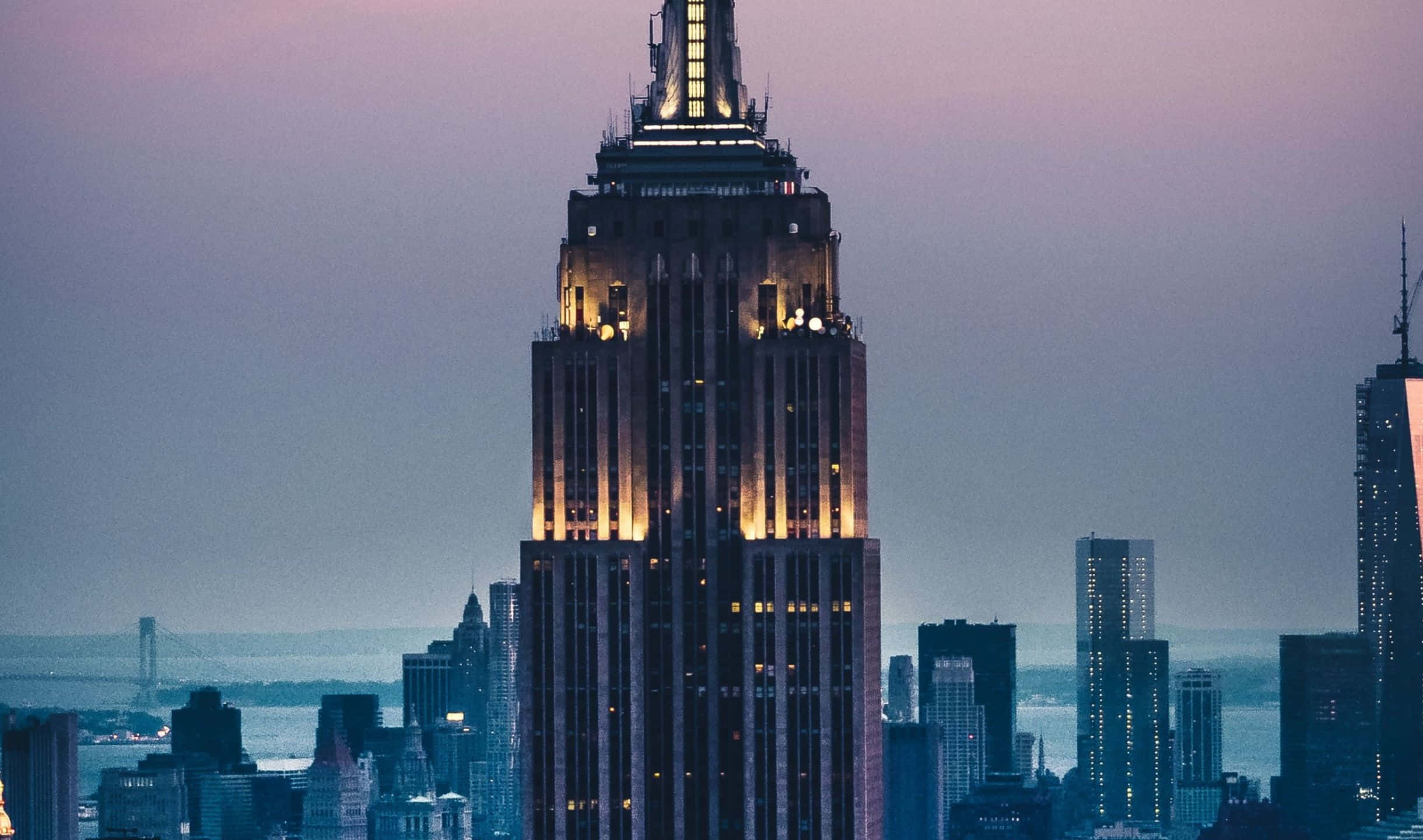 2440x1440 Empire State Building 桌布 壁紙 — 帝國大廈在昏暗的紫色天空下溫暖地發光，城市的燈光開始在下方閃爍。