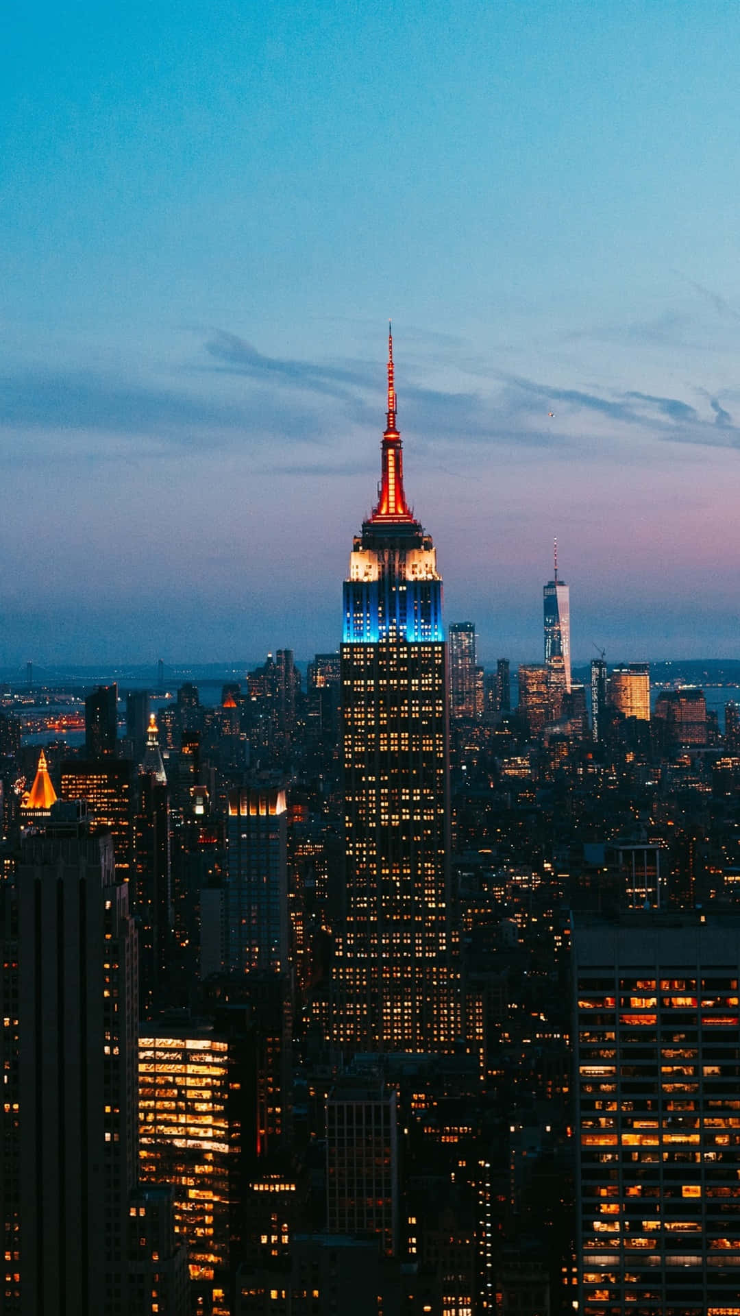 New York 桌布 壁紙 — 帝國大廈在暮色天空中，在密集的紐約市天際線上方閃爍著藍色和橙色的燈光。