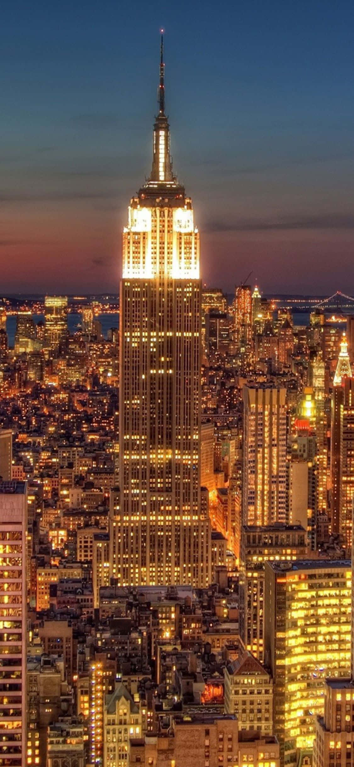 iPhone Xs Ny City 桌布 壁紙 — 帝國大廈在暮色天空中閃耀金色光芒，紐約市的燈光在下方閃爍。