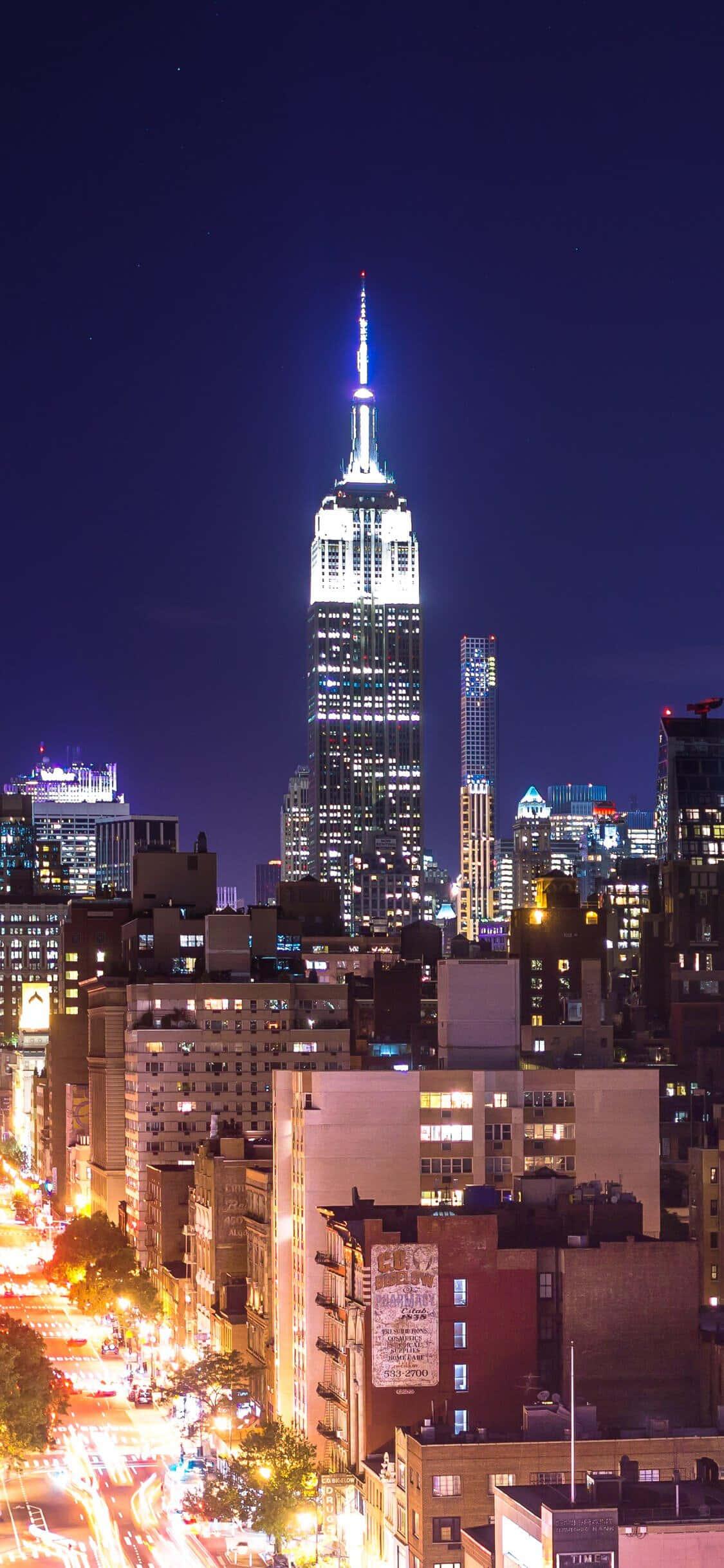New York City Night iPhone 桌布 壁紙 — 帝國大廈在深邃的夜空中閃耀著光芒，聳立在充滿活力的城市景觀之上，下方繁忙街道的光線條紋清晰可見。