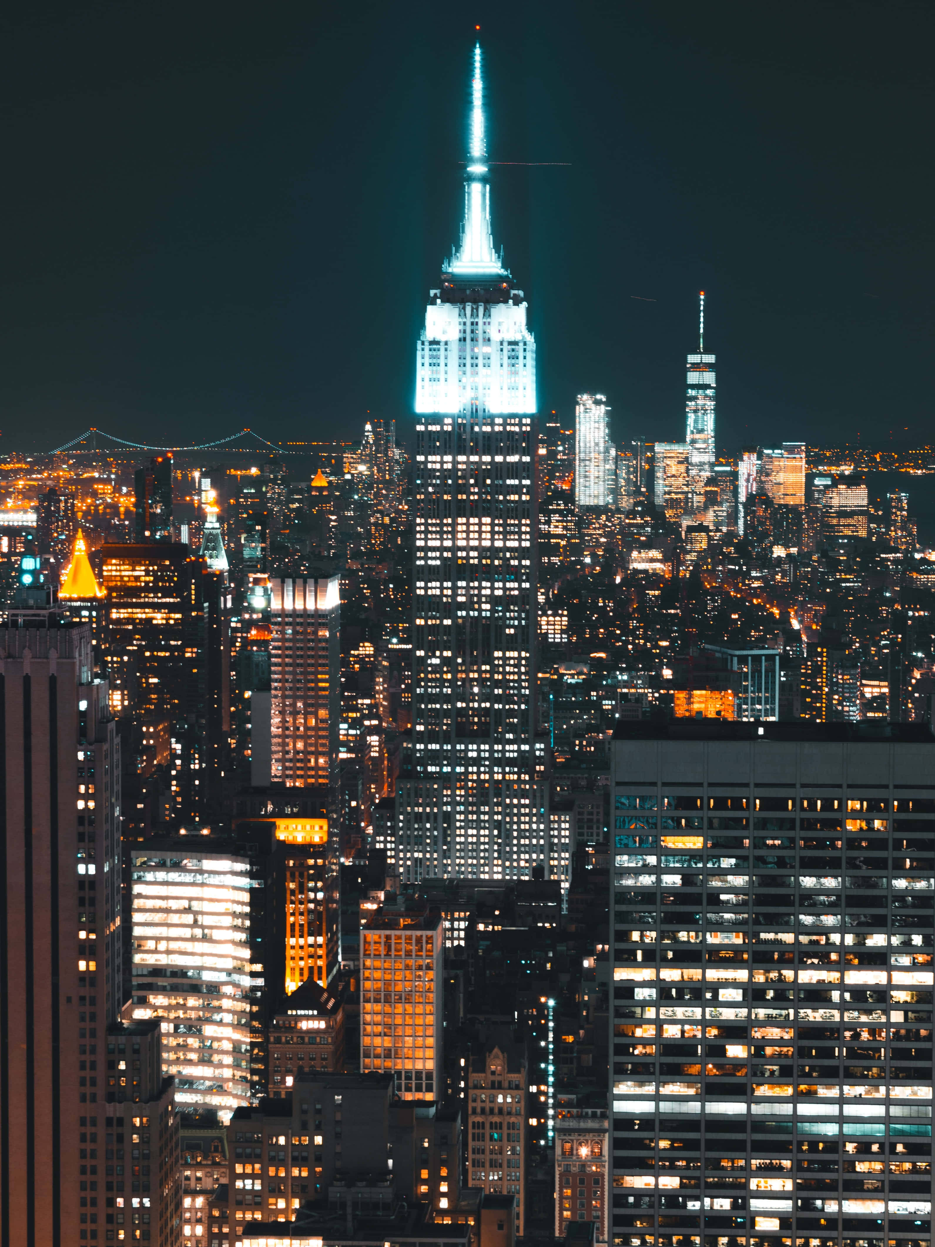 New York State 桌布 壁紙 — 帝國大廈在漆黑的夜空下閃耀著藍色光芒的壯麗夜景，四周環繞著密集的都市天際線，盡是燈火通明的摩天大樓。