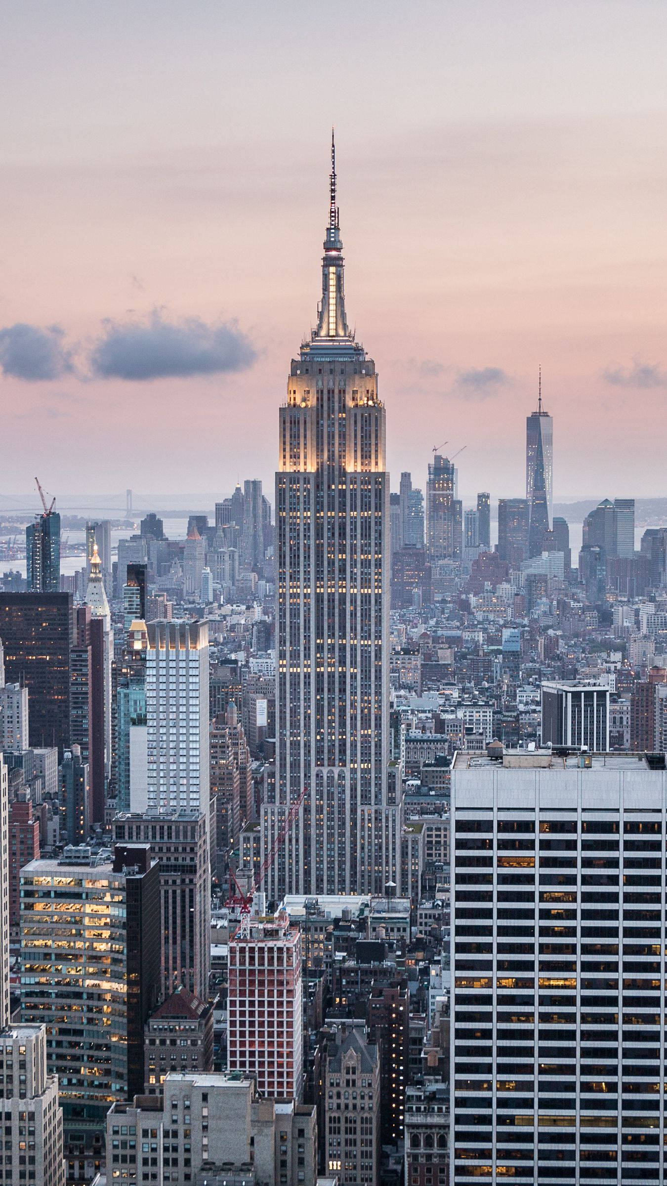 new york city iPhone x 桌布 壁紙 — 帝國大廈在紐約市密集的天際線上方，襯著柔和的粉紅與橙色暮色天空，亮起燈光矗立。