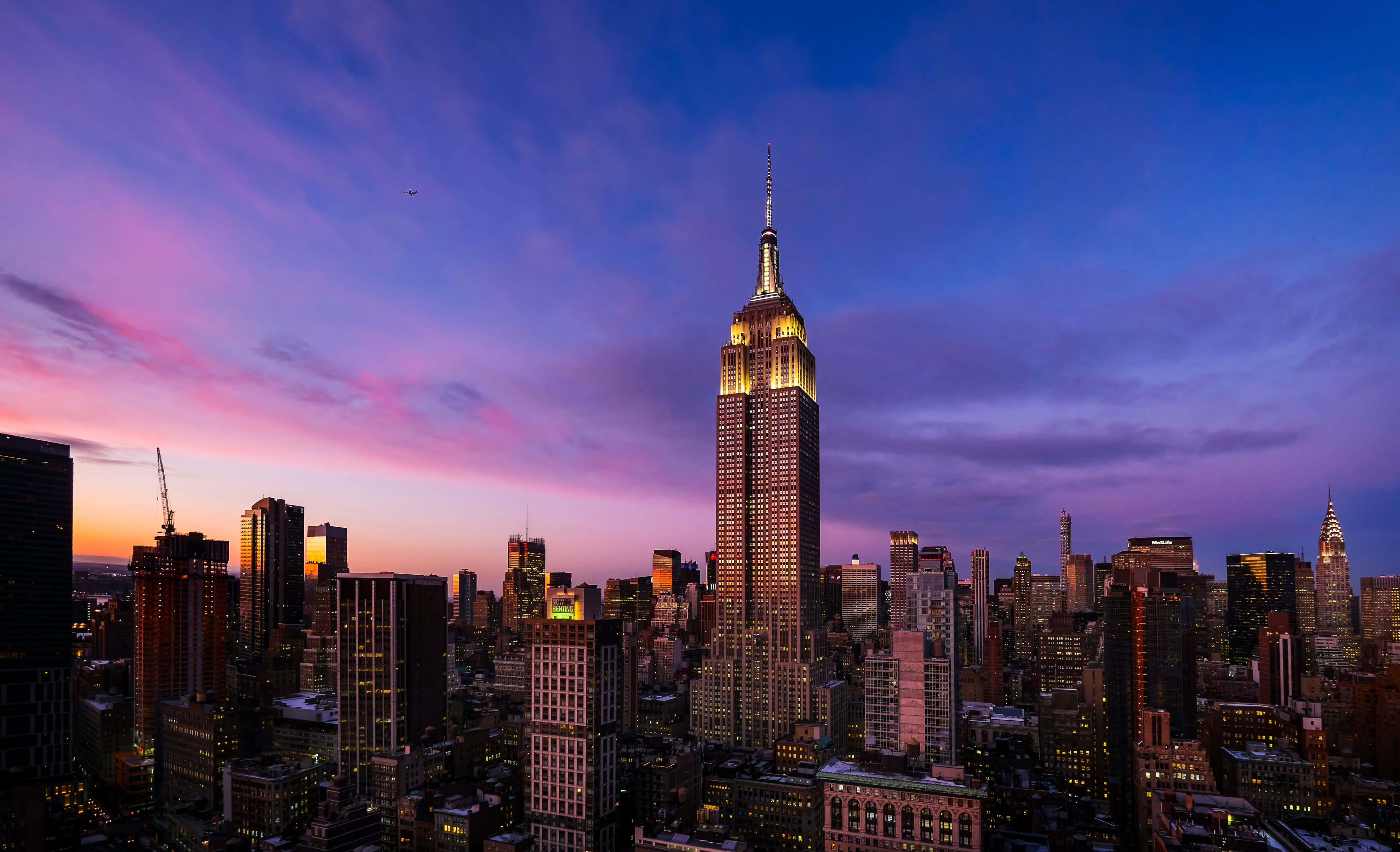 Empire State Building 桌布 壁紙 — 帝國大廈在紐約市密集的天際線上方，閃耀著金色光芒，映襯著充滿活力的紫色和粉紅色暮色天空。