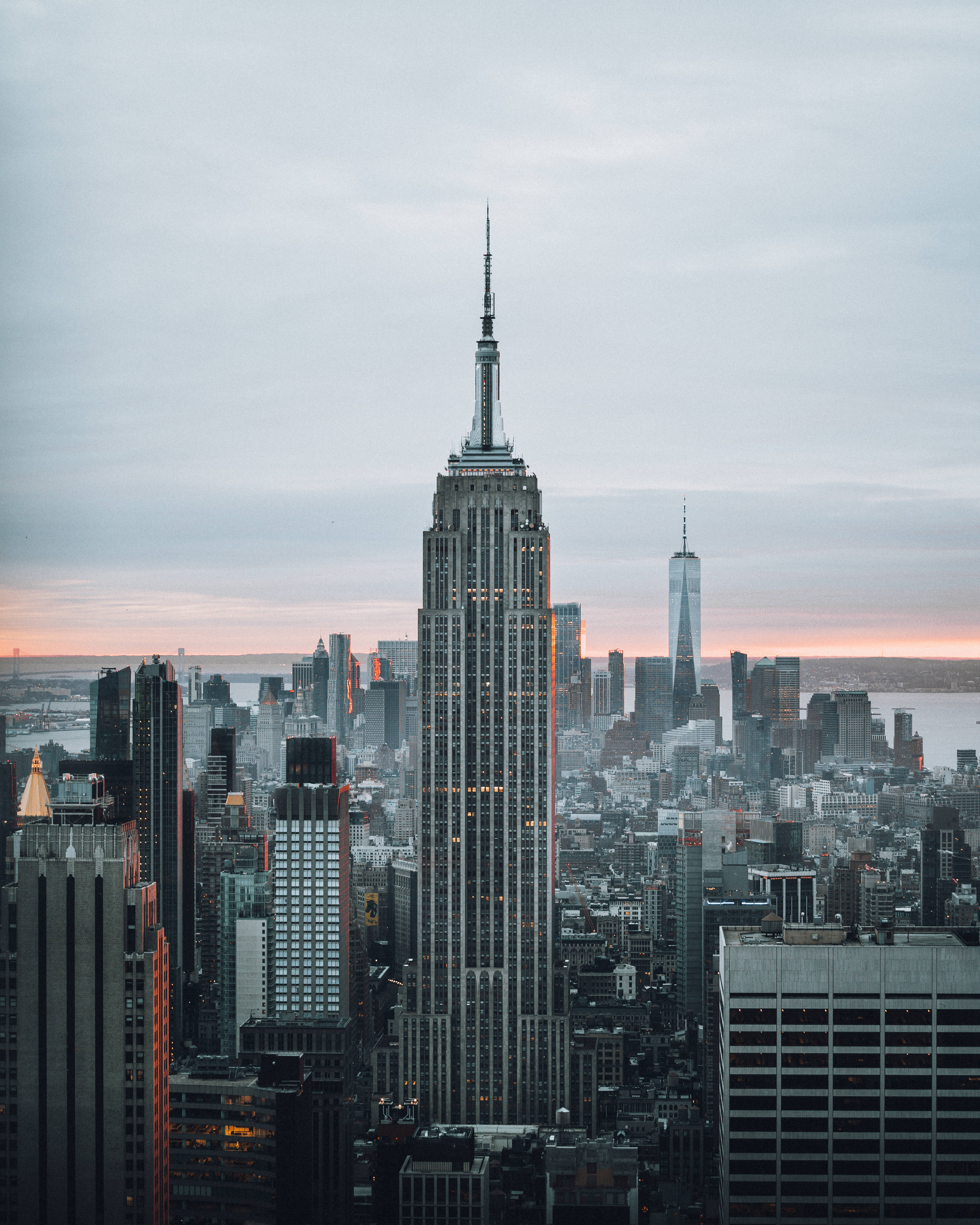 empire state 桌布 壁紙 — 帝國大廈在黃昏時分聳立在密集的紐約市天際線之上，上方有柔和的粉紅色和灰色雲彩。
