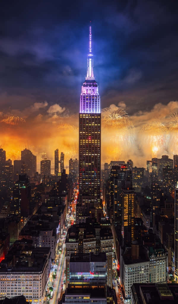 New York City Night iPhone 桌布 壁紙 — 帝國大廈在黑暗的天空中發出紫色和藍色的光芒，煙火在紐約市上空以金色爆發綻放。