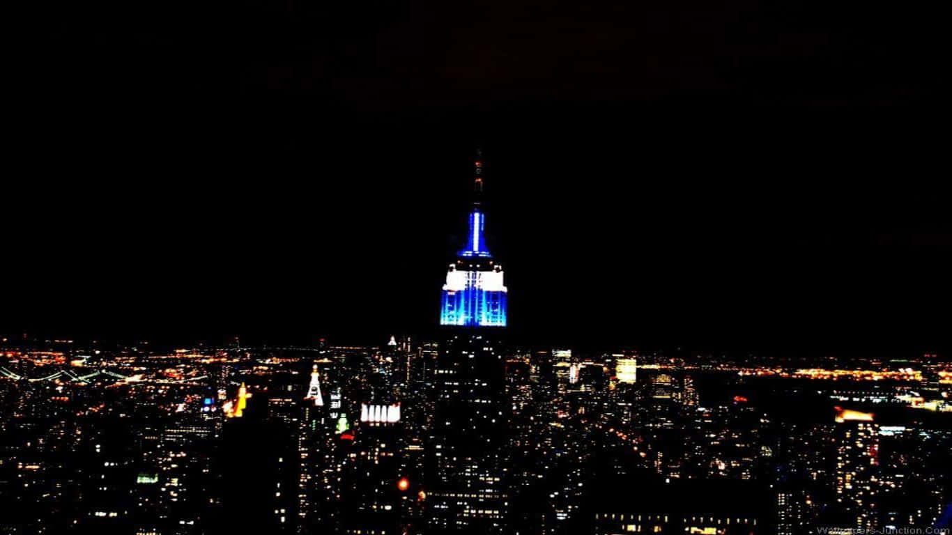 1366x768 Empire State Building 桌布 壁紙 — 帝國大廈在黑色天空下發出藍光，高聳於夜晚閃爍燈光的廣闊城市景觀之上。
