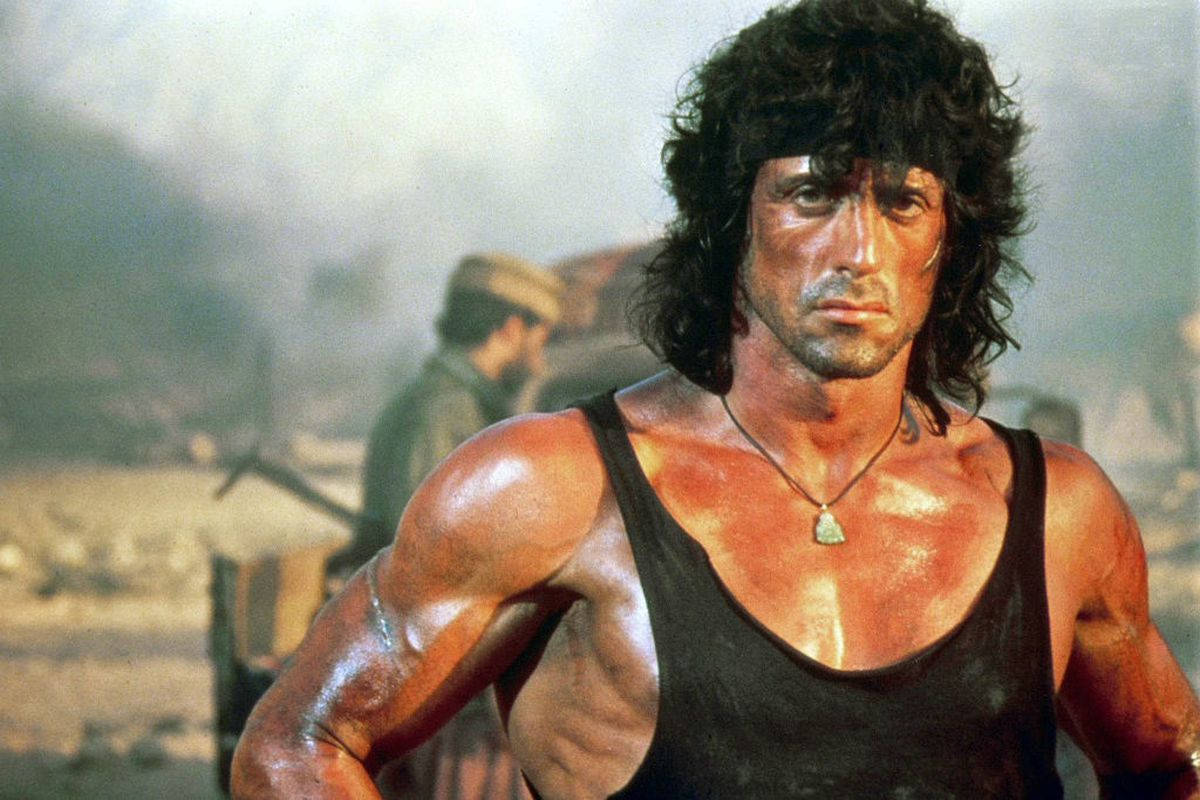 rambo 桌布 壁紙 — 席維斯史特龍飾演的藍波在塵土飛揚的戰區中，背景有煙霧和士兵，他凝視著前方。