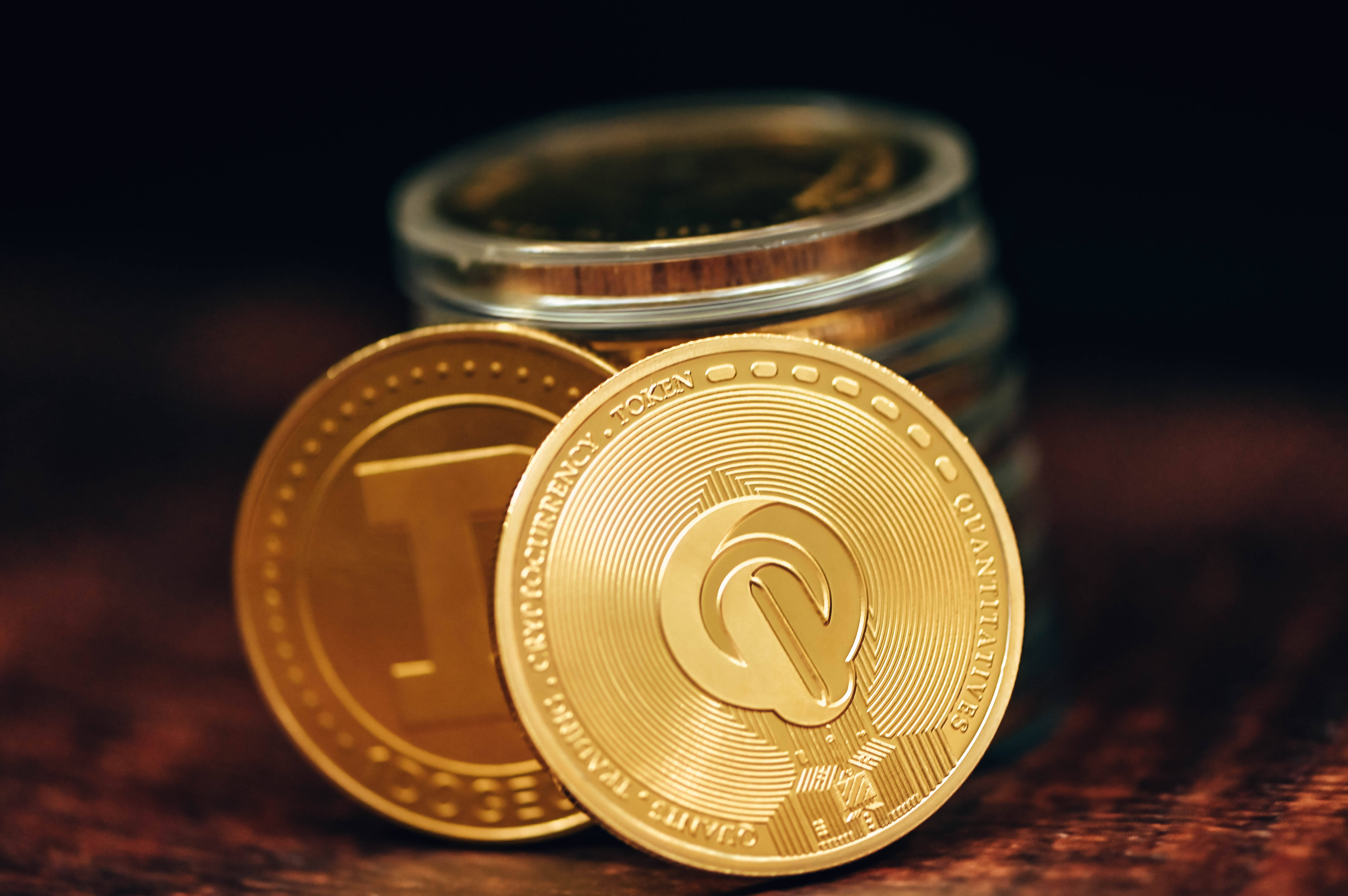 金融 桌布 壁紙 — 帶有「Quantum」標誌的金色加密貨幣硬幣放在木質表面上，旁邊有一個裝滿更多硬幣的玻璃罐。