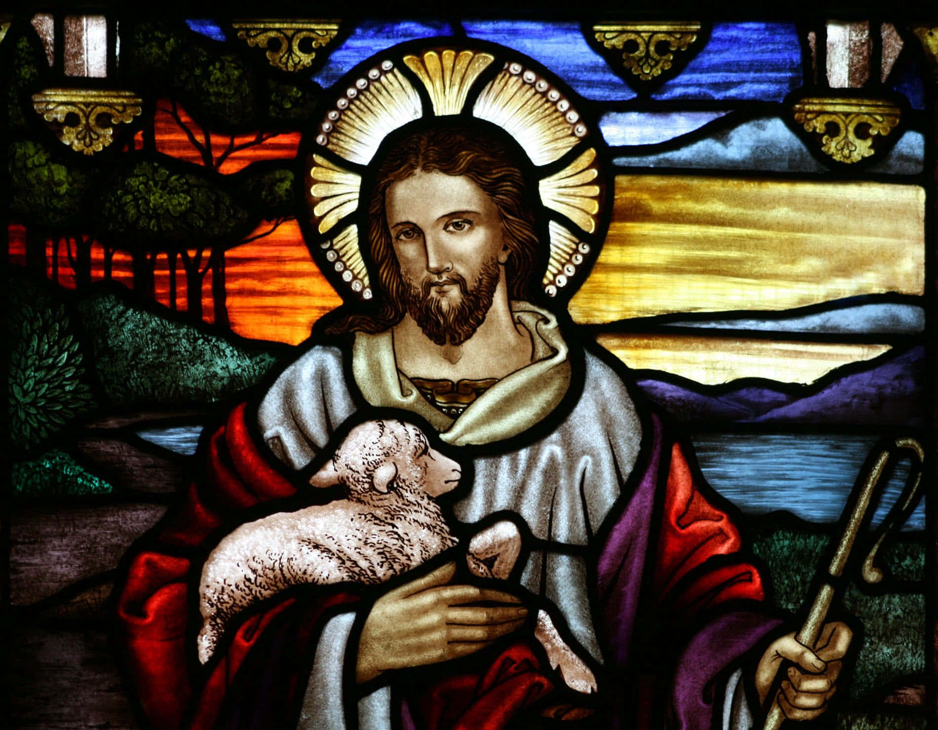 Jesus With Sheep 桌布 壁紙 — 帶有光環的耶穌基督在樹木和日落山丘的彩繪玻璃背景下抱著一隻羔羊和權杖。