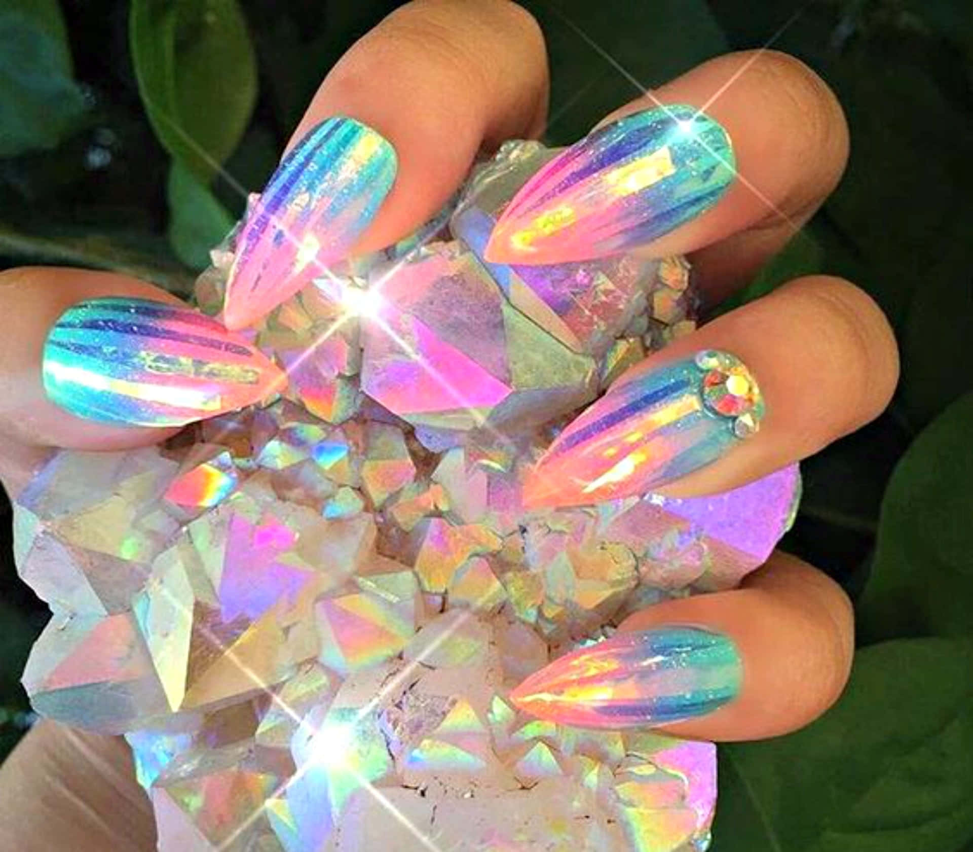 Cute Nails 桌布 壁紙 — 帶有彩虹色指甲的手，在茂密的綠葉中捧著閃閃發光的水晶簇。
