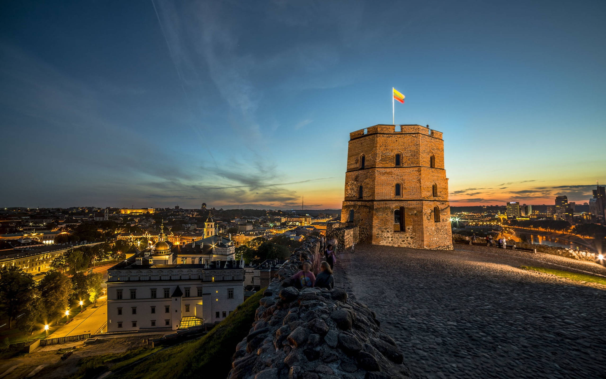Vilnius 桌布 壁紙 — 帶有旗幟的古老石塔俯瞰著暮色中閃爍的城市景觀，天空帶有條紋。