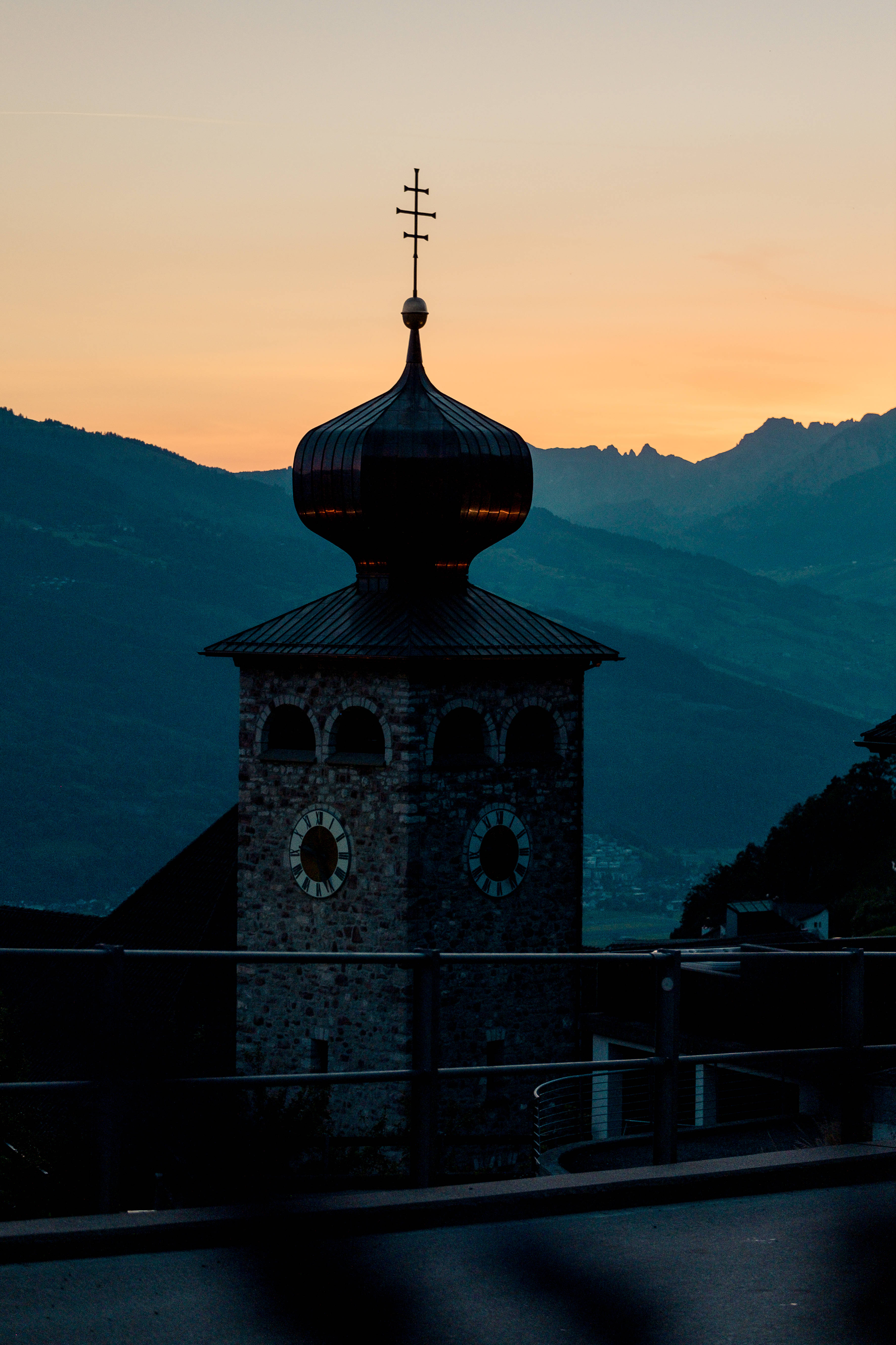 Liechtenstein 桌布 壁紙 — 帶有洋蔥形圓頂和十字架的教堂尖塔剪影，在遠山之上溫暖的日落天空中矗立。