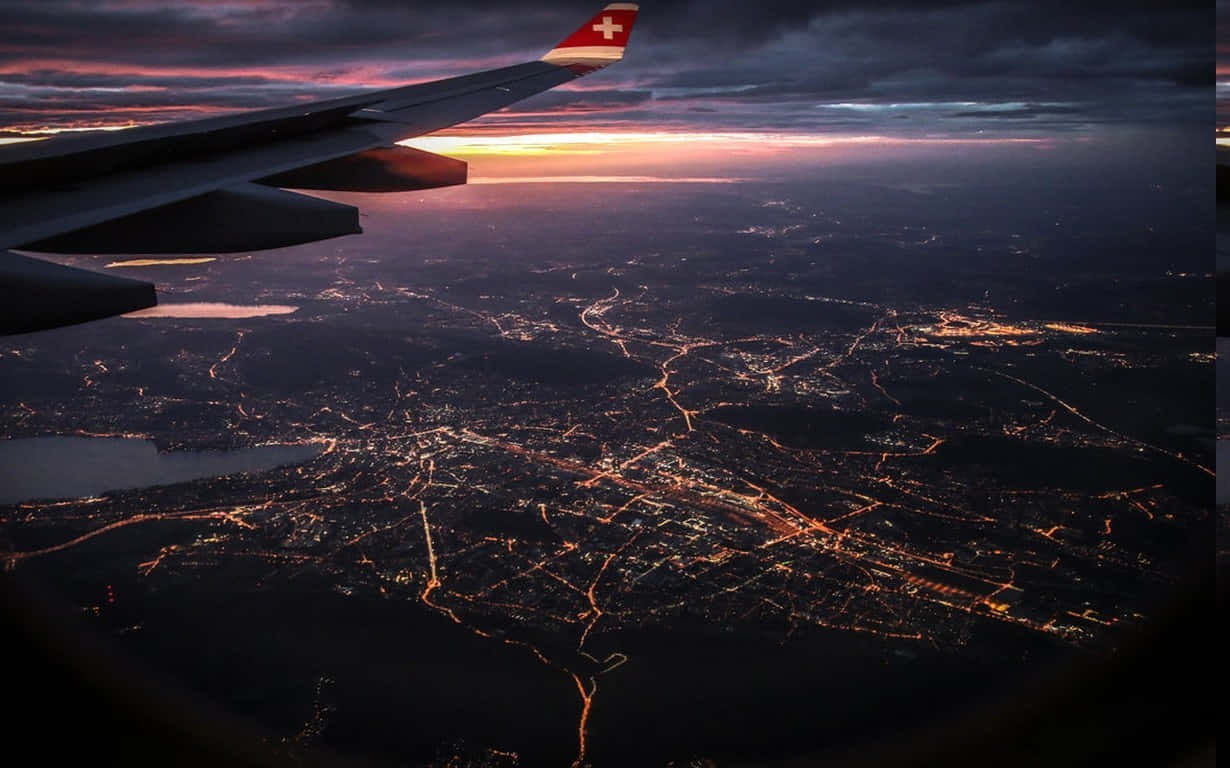 Airplane Window 桌布 壁紙 — 帶有瑞士國旗的飛機機翼俯瞰著廣闊的城市景觀，在黃昏時被金色的燈光照亮。